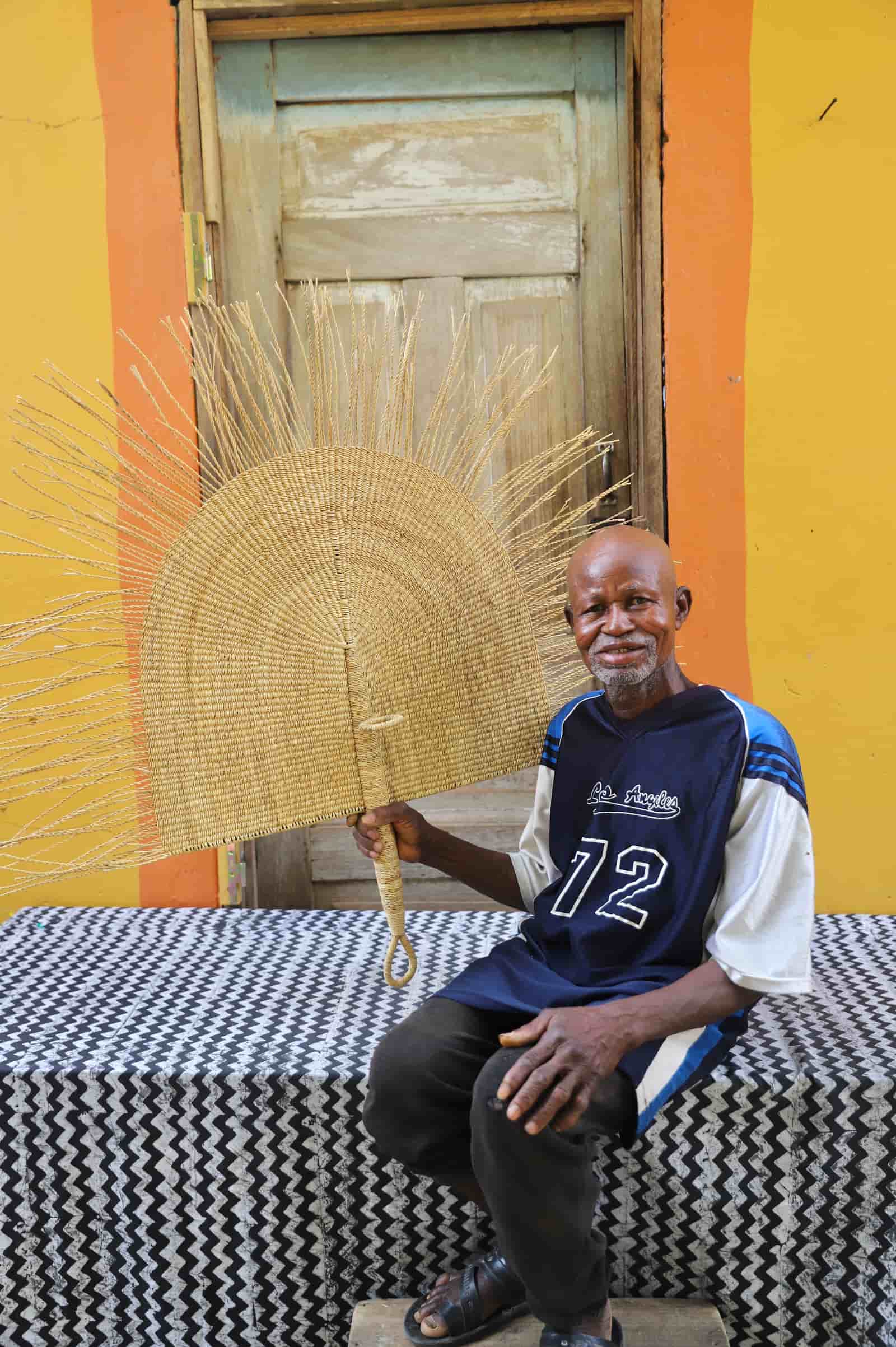 Man holding a woven fan in front of a colorful wall with a wooden door.