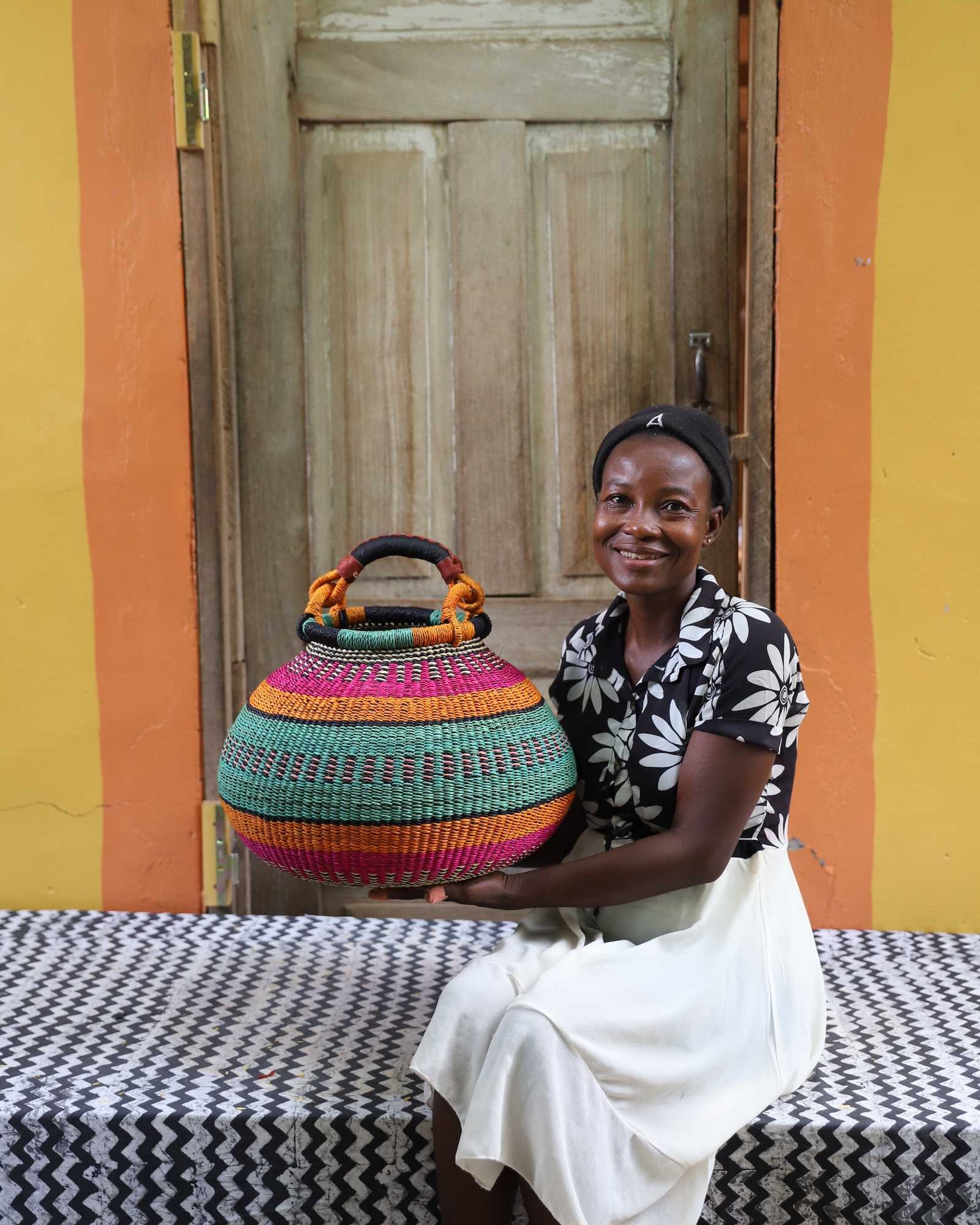 Large handwoven Bolga pot basket in bold bands of pink, teal and amber, held by a smiling woman in Bolgatanga.
