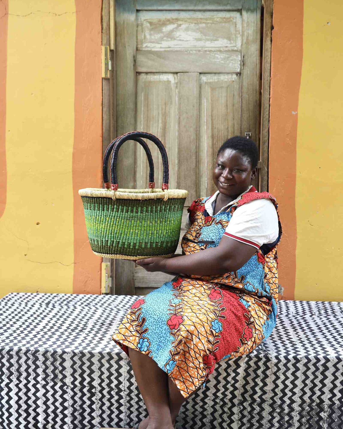 Small oval Bolga shopping basket in natural with green bands, held by a seated woman in red.