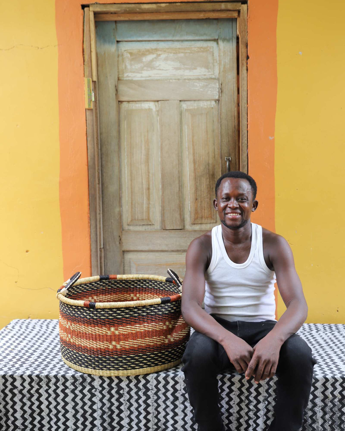 Large handwoven Bolga storage tub in natural straw with orange and black diamonds, beside a seated man.