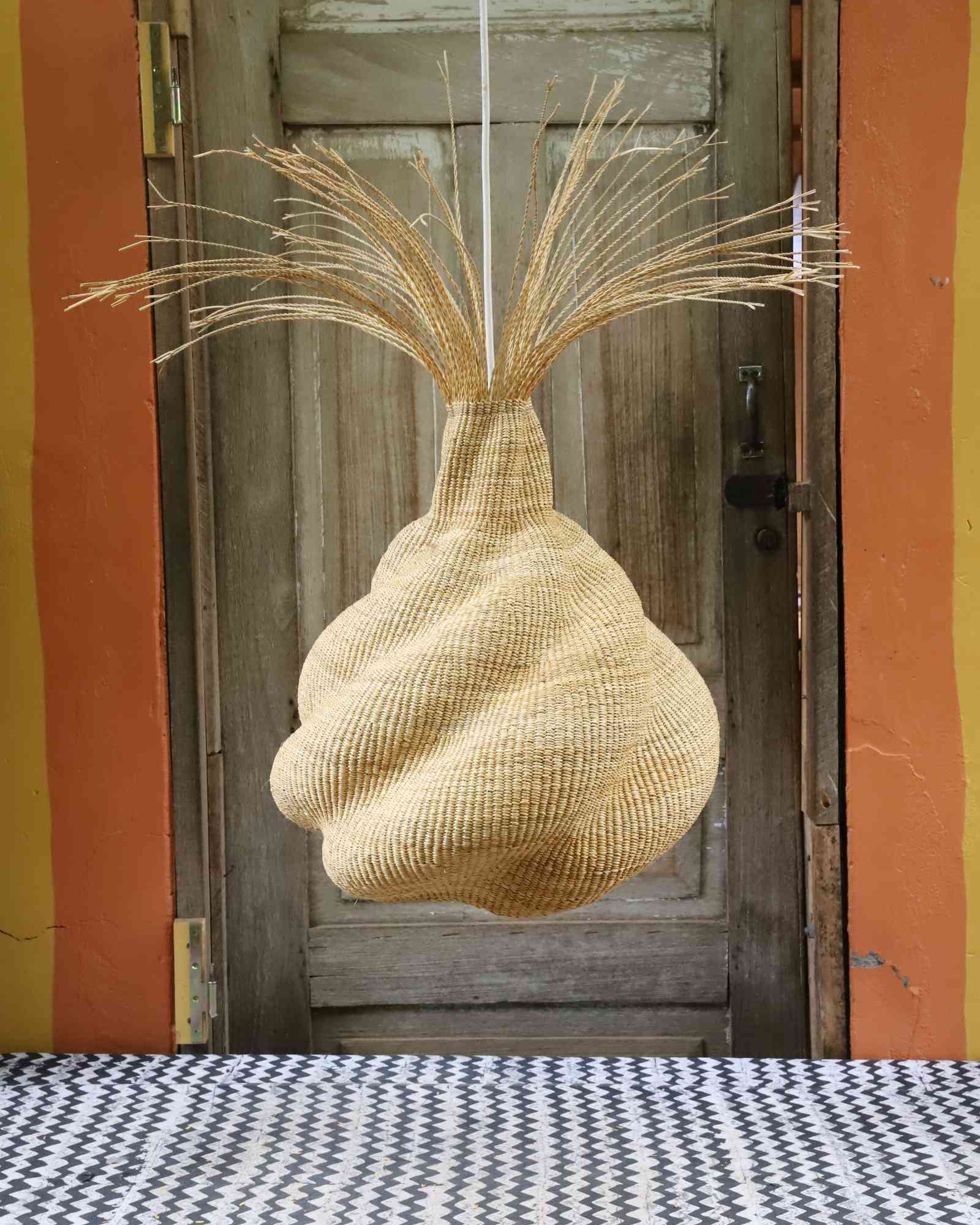 Helix pendant lampshade with a twisted woven form and fine straw fringe, hung against a weathered door.