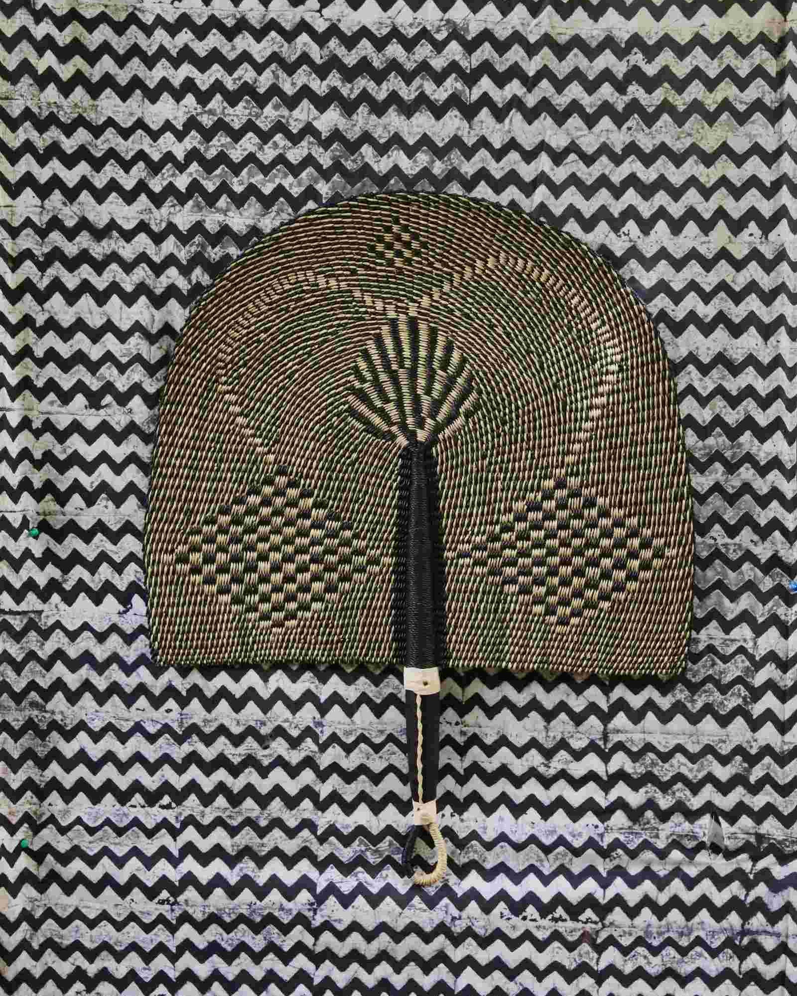 Naba Penga handwoven wall fan in natural straw and black geometric check pattern, on a chevron-print backdrop.