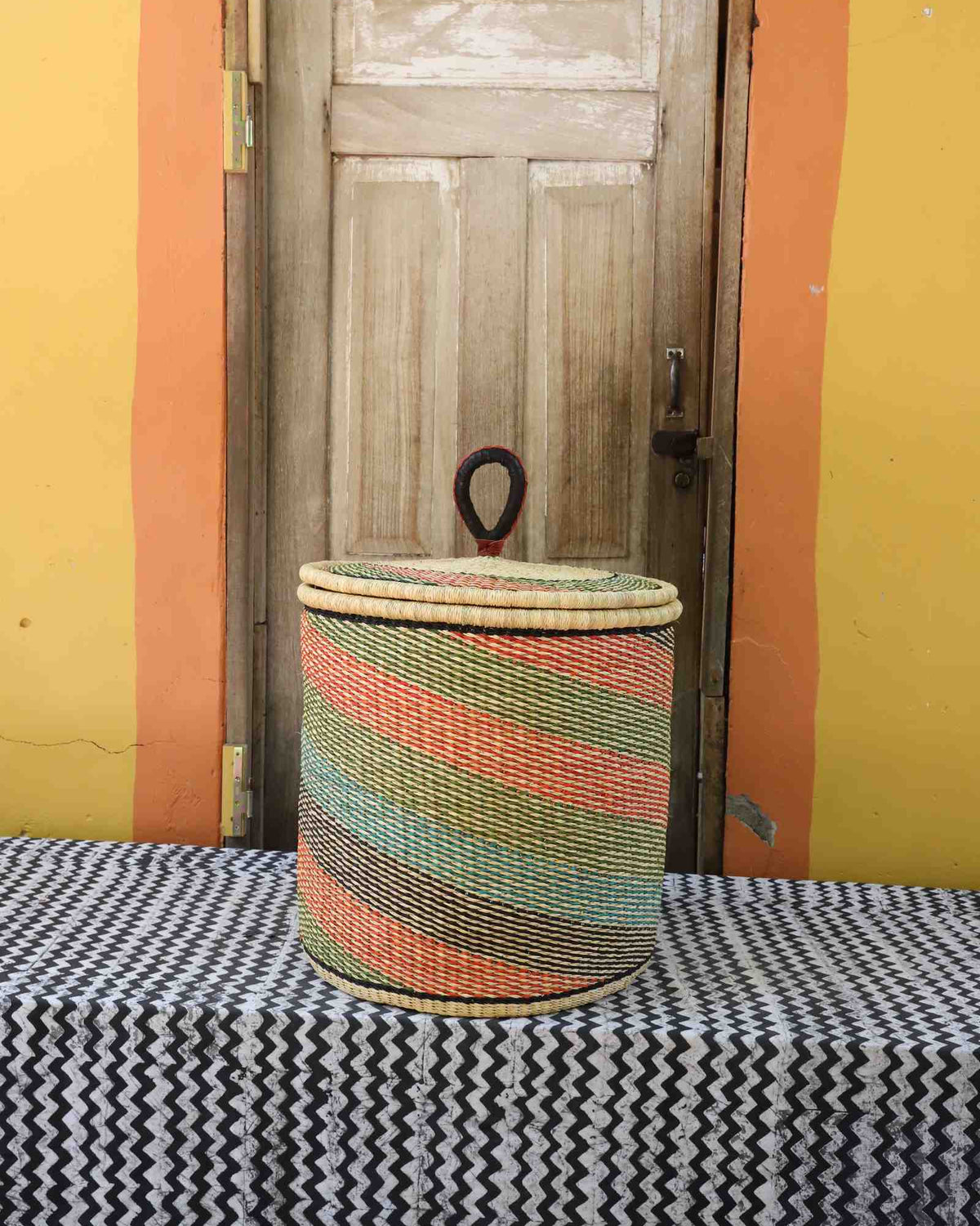 Handwoven Bolga laundry basket with coiled rim and lid in natural straw with fine pink, green and orange diagonal stripes.