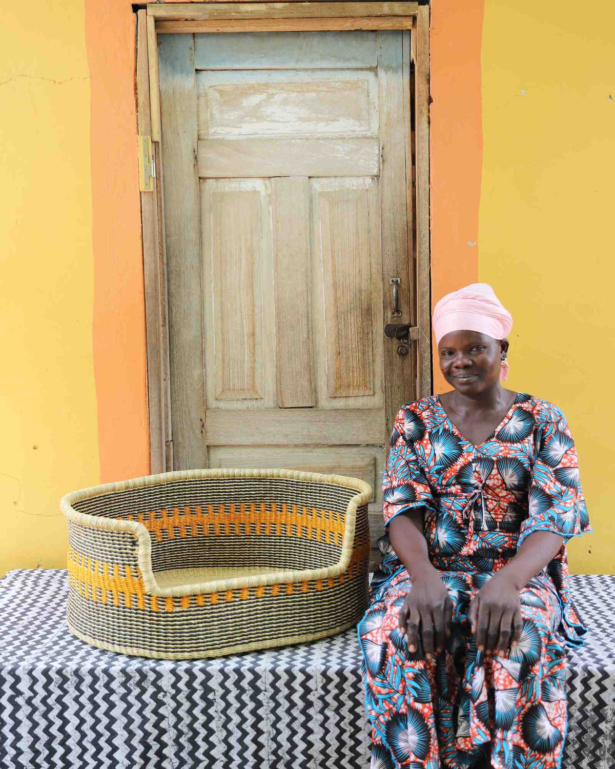 Large handwoven Bolga pet bed in natural straw with fine orange stripes, beside a seated woman in a printed dress.