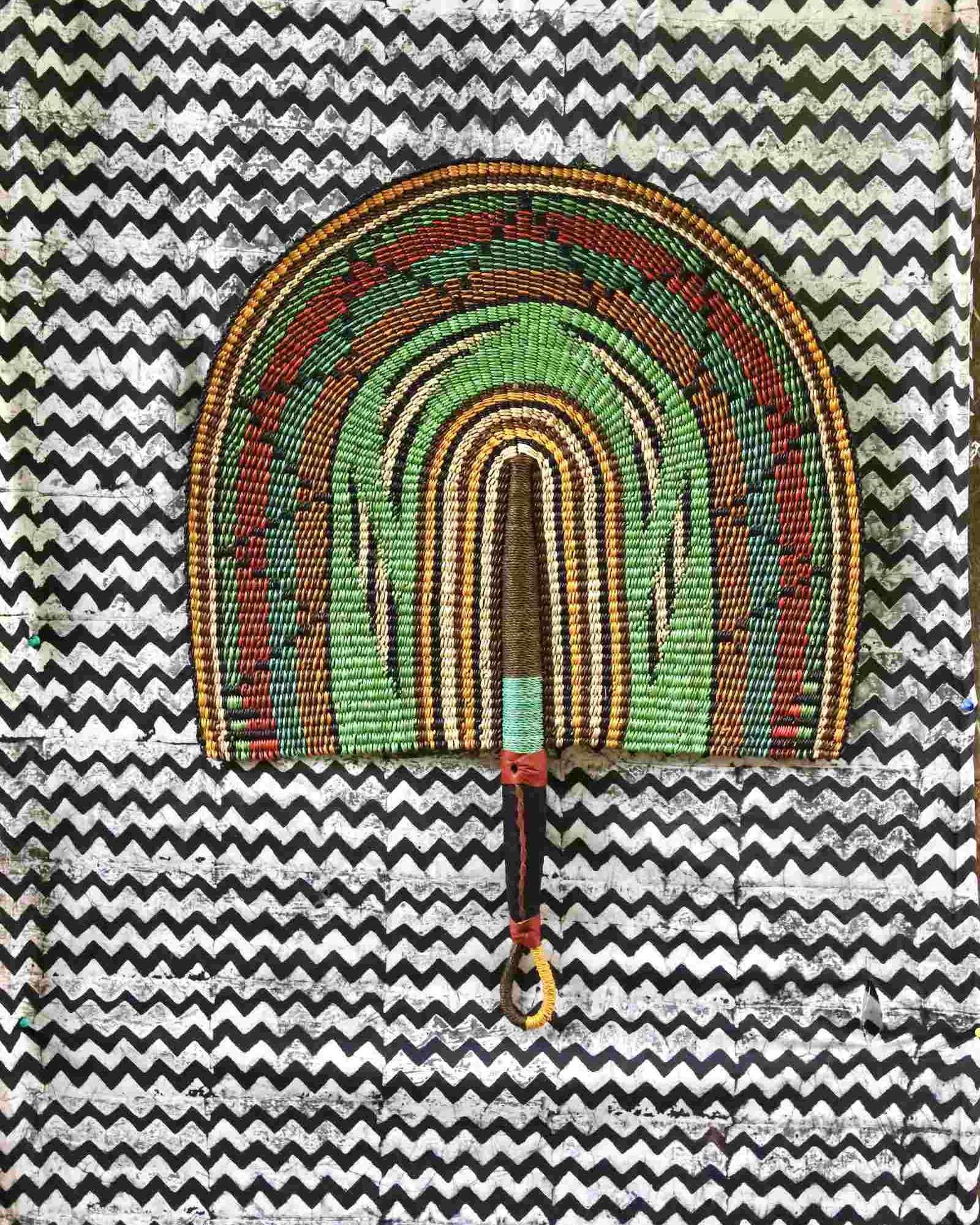 Naba Penga handwoven wall fan with arch bands in green, terracotta and amber, on a chevron-print fabric backdrop.