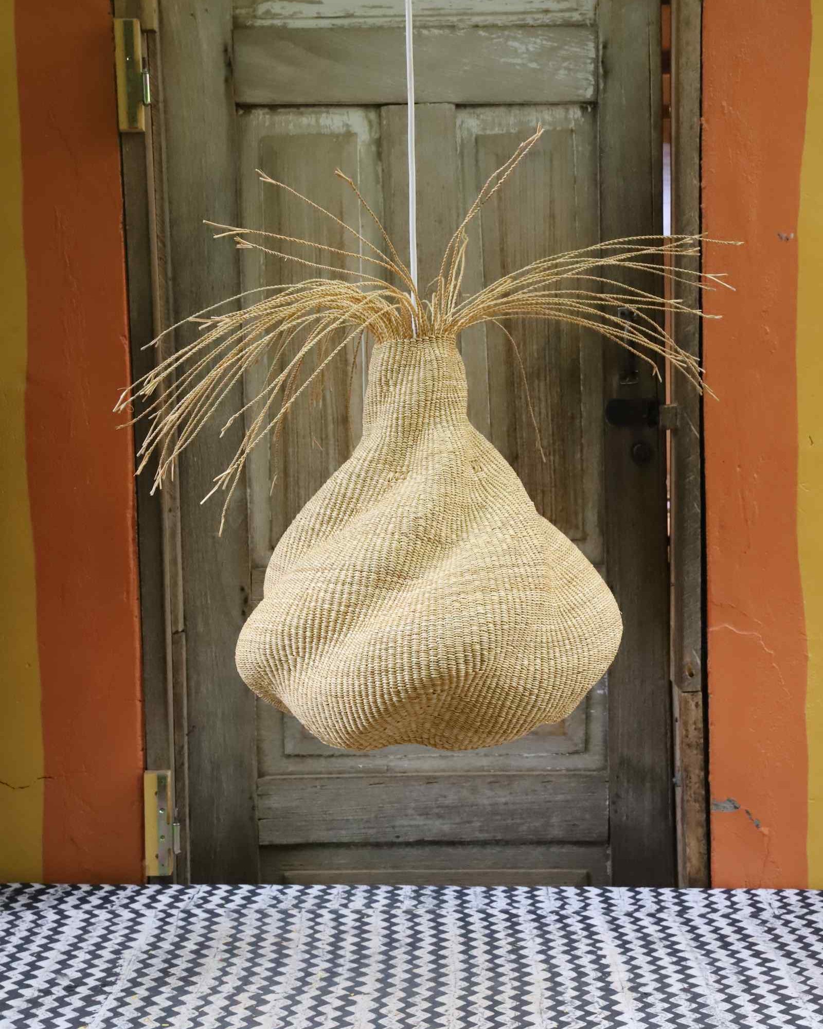 Twisted Helix pendant lampshade in woven elephant grass with straw fringe, hung against a wooden door.
