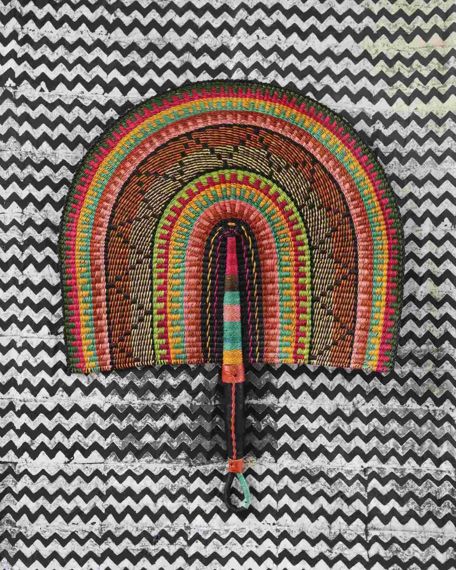 Naba Penga wall fan in natural straw with black, green and orange banding, laid on a chevron cloth.