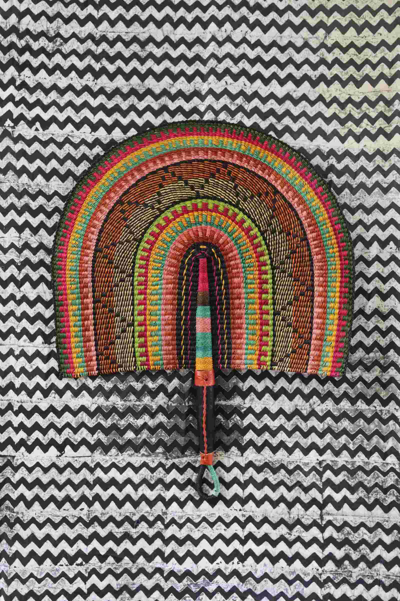 Naba Penga wall fan in natural straw with black, green and orange banding, laid on a chevron cloth.