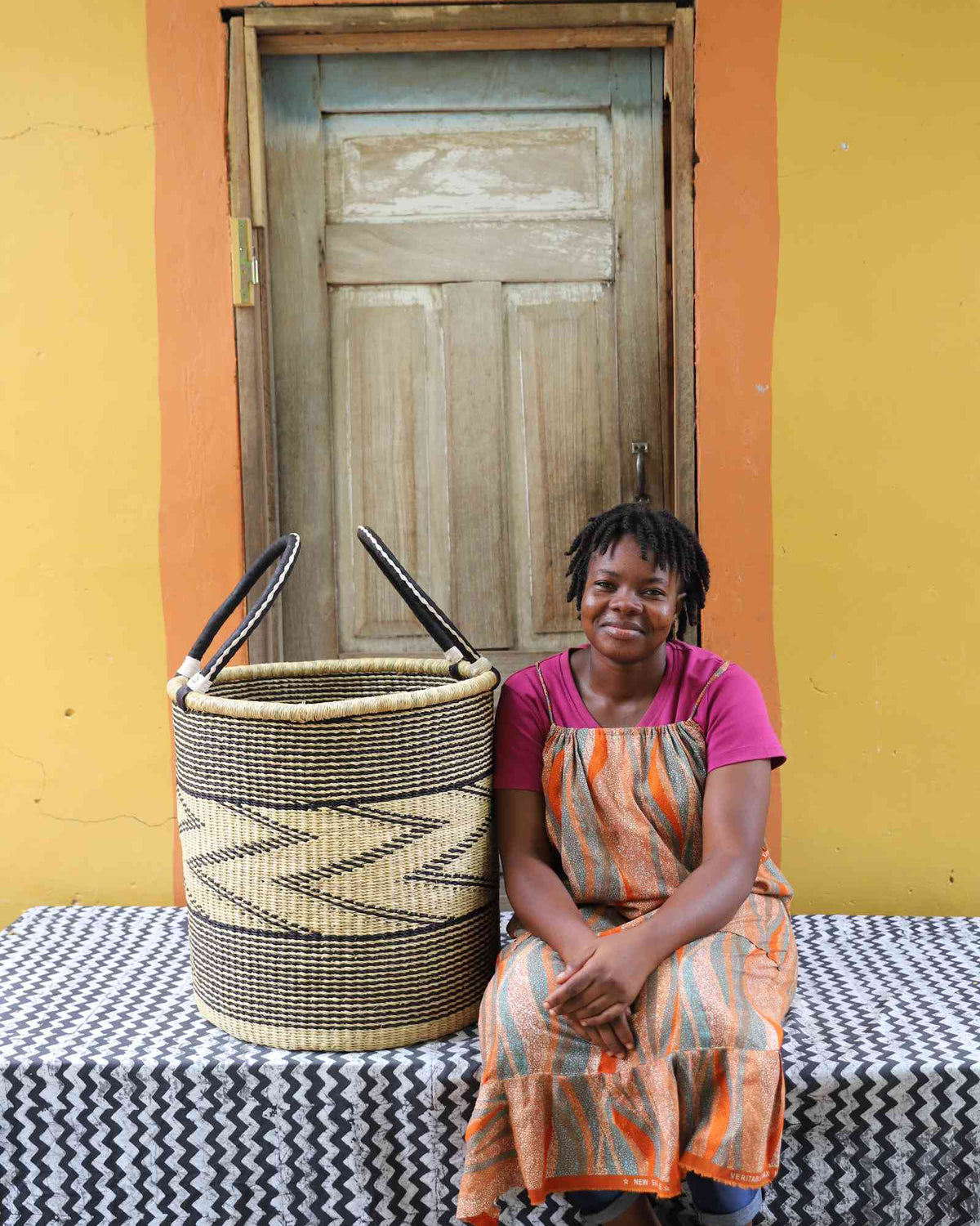 Large handwoven Bolga laundry basket in natural with fine black zigzag wave, beside a seated woman in orange.