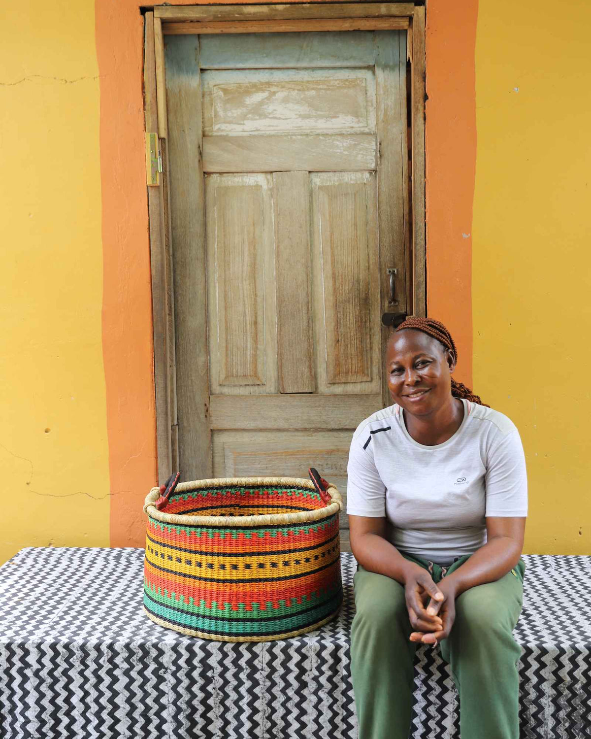 Medium handwoven Bolga storage tub in green with orange and yellow bands, beside a seated woman in white.
