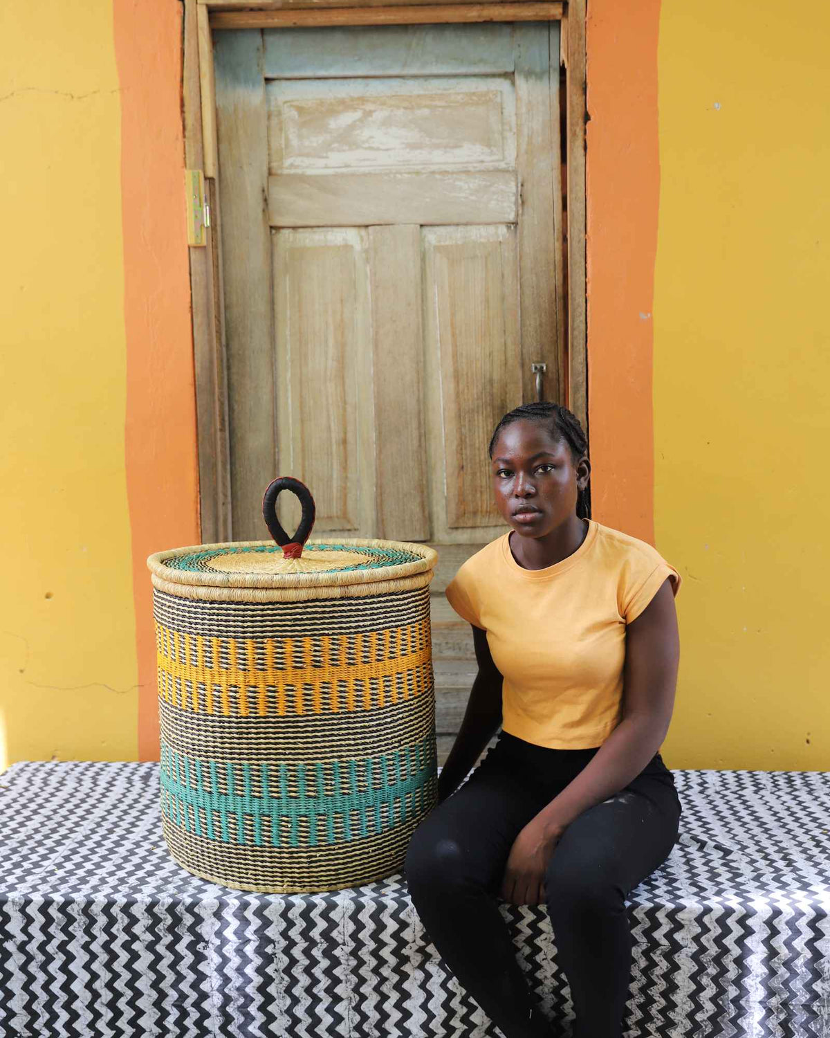 Handwoven Bolga laundry basket in natural straw with yellow and teal bands, beside a seated woman in yellow.