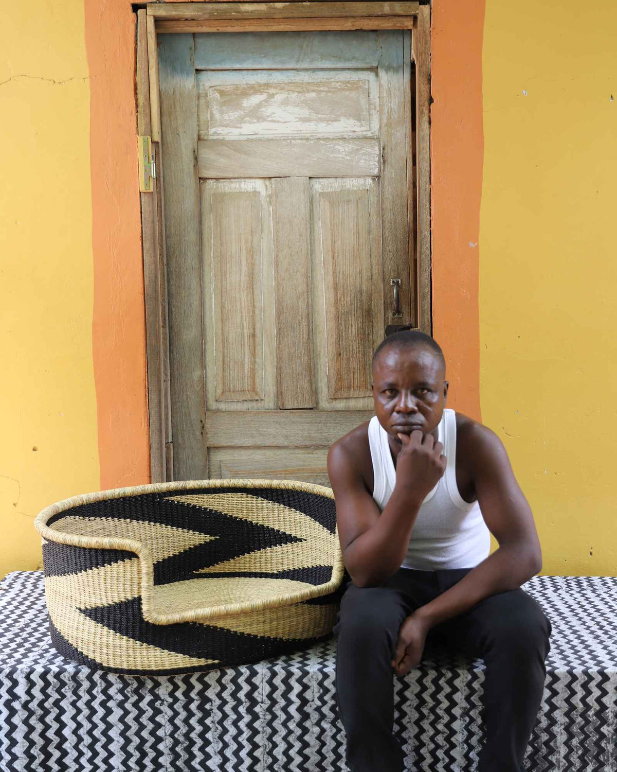 Large handwoven Bolga pet bed in natural straw with bold black chevron, beside a seated man in a white vest.