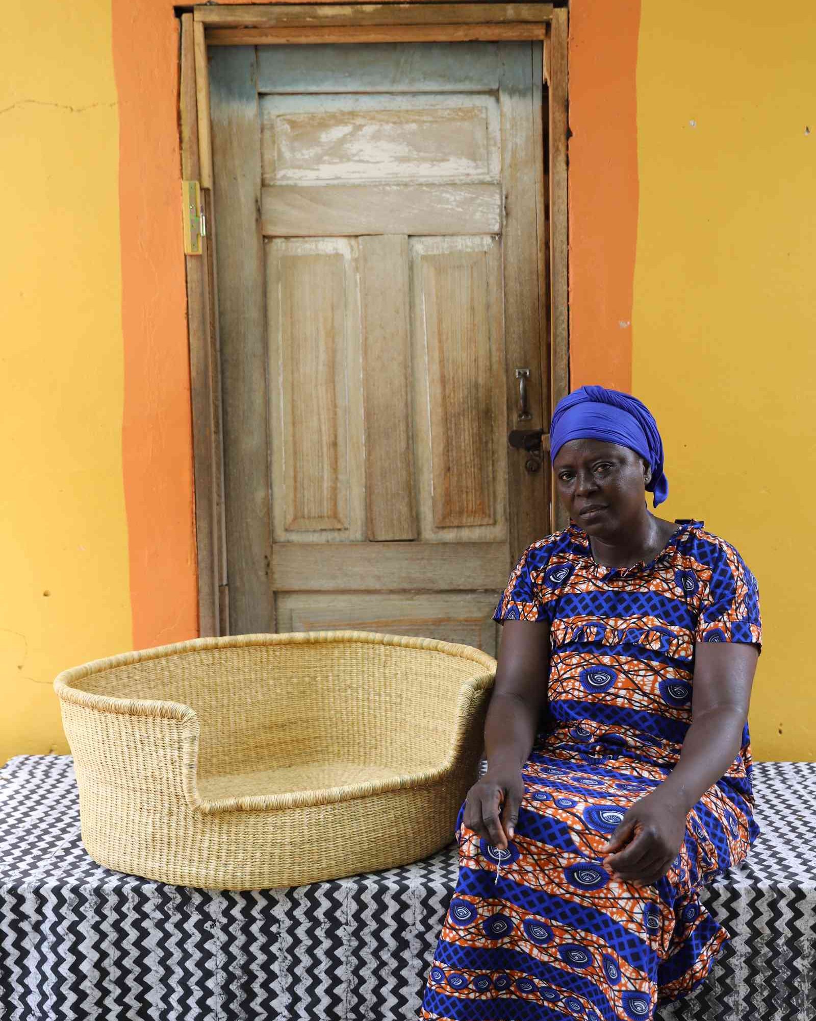 Large handwoven Bolga pet bed in plain natural straw, beside a seated woman in a printed dress.