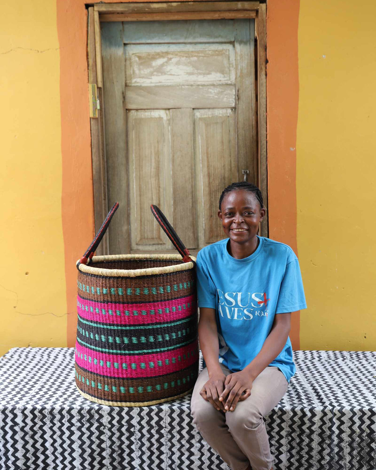 Large handwoven Bolga laundry basket in natural with pink and dark bands, beside a seated woman in blue.