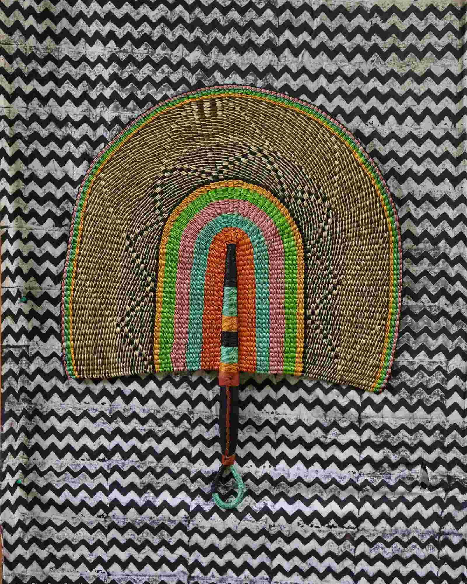 Naba Penga handwoven wall fan with natural straw outer arch and inner bands of pink, teal and orange, on a chevron backdrop.