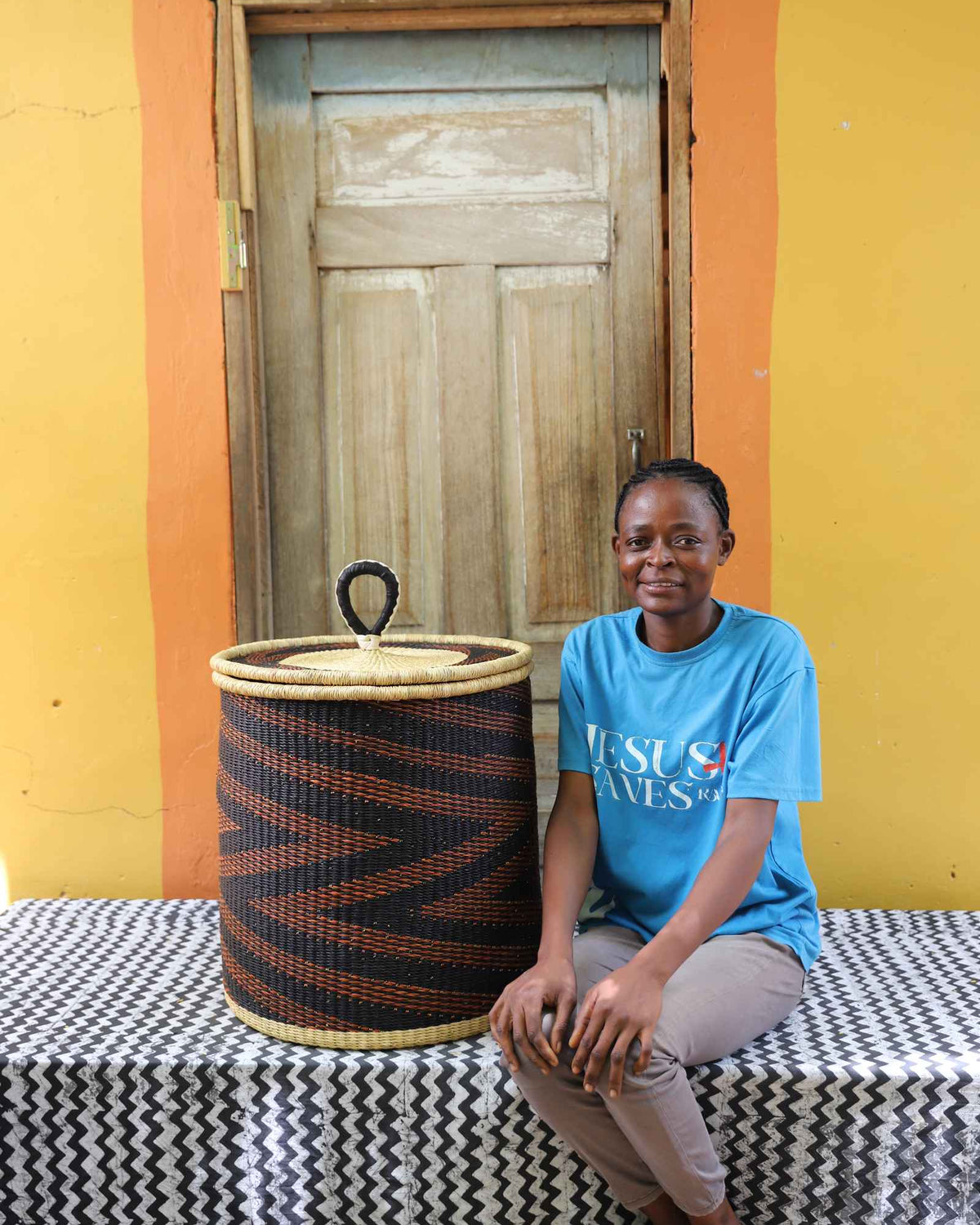 Handwoven Bolga laundry basket in dark navy with rust-orange waves, beside a seated woman in blue.