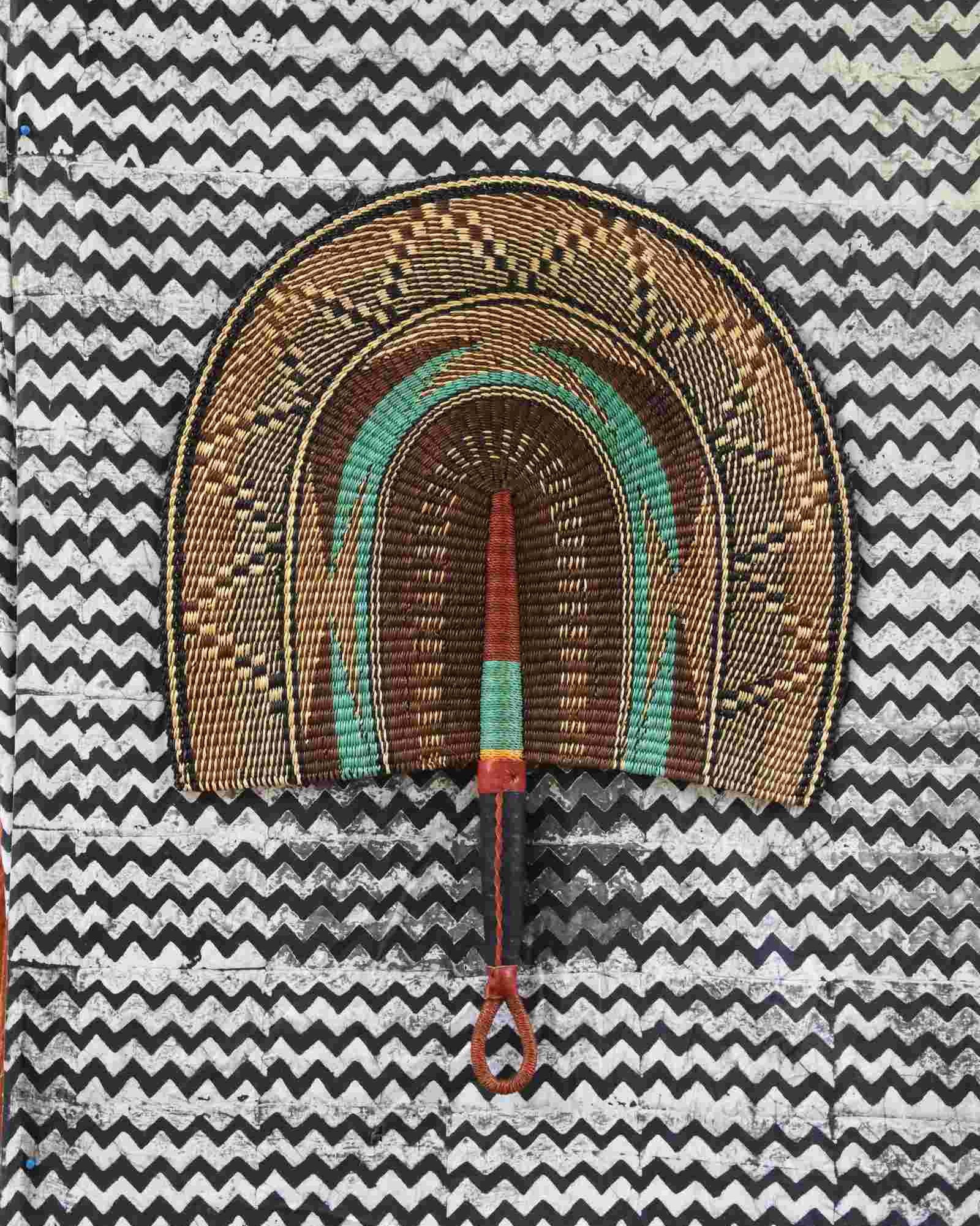 Naba Penga wall fan in natural straw with a teal inner arch and dark patterned border, laid on a chevron cloth.