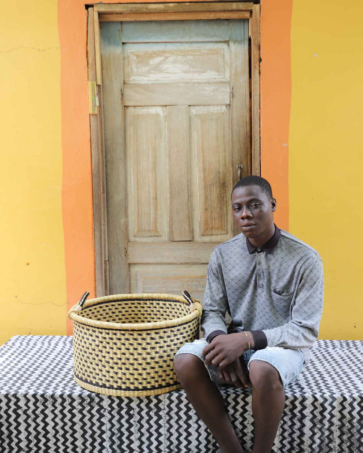 Medium handwoven Bolga storage tub in plain natural straw with dark handles, beside a seated man.