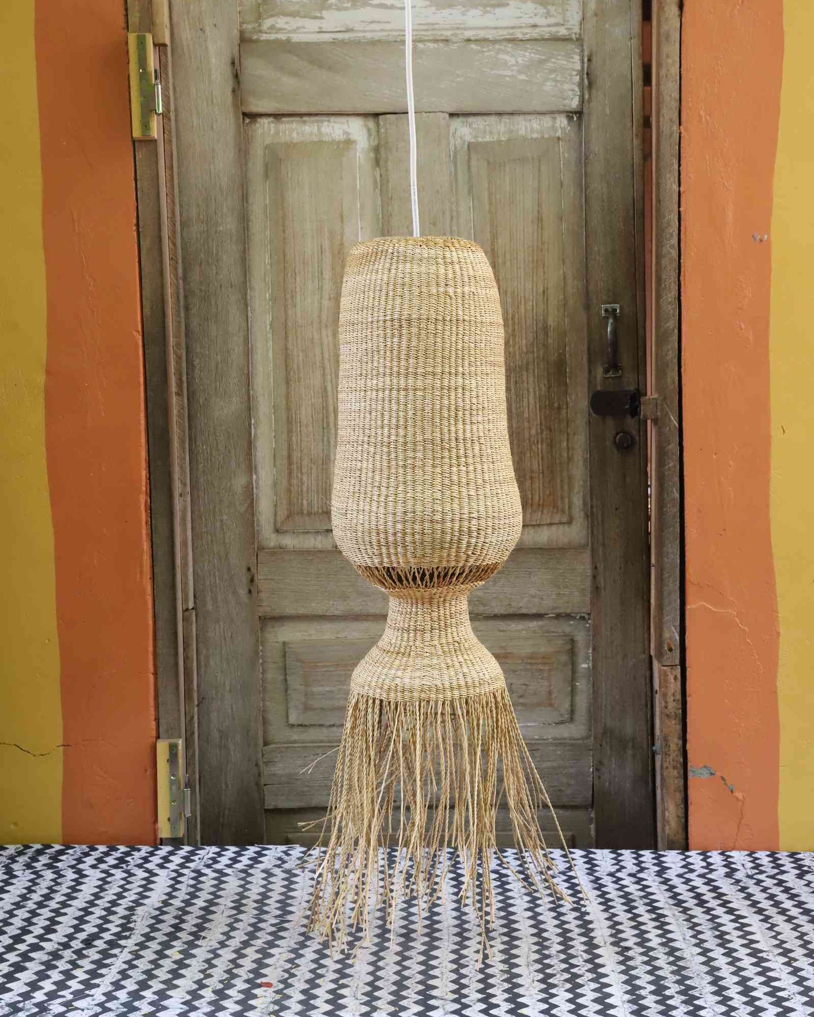 Elephant grass Abyssal pendant lampshade with a bulbed waist and neat fringe, hung against a wooden door.