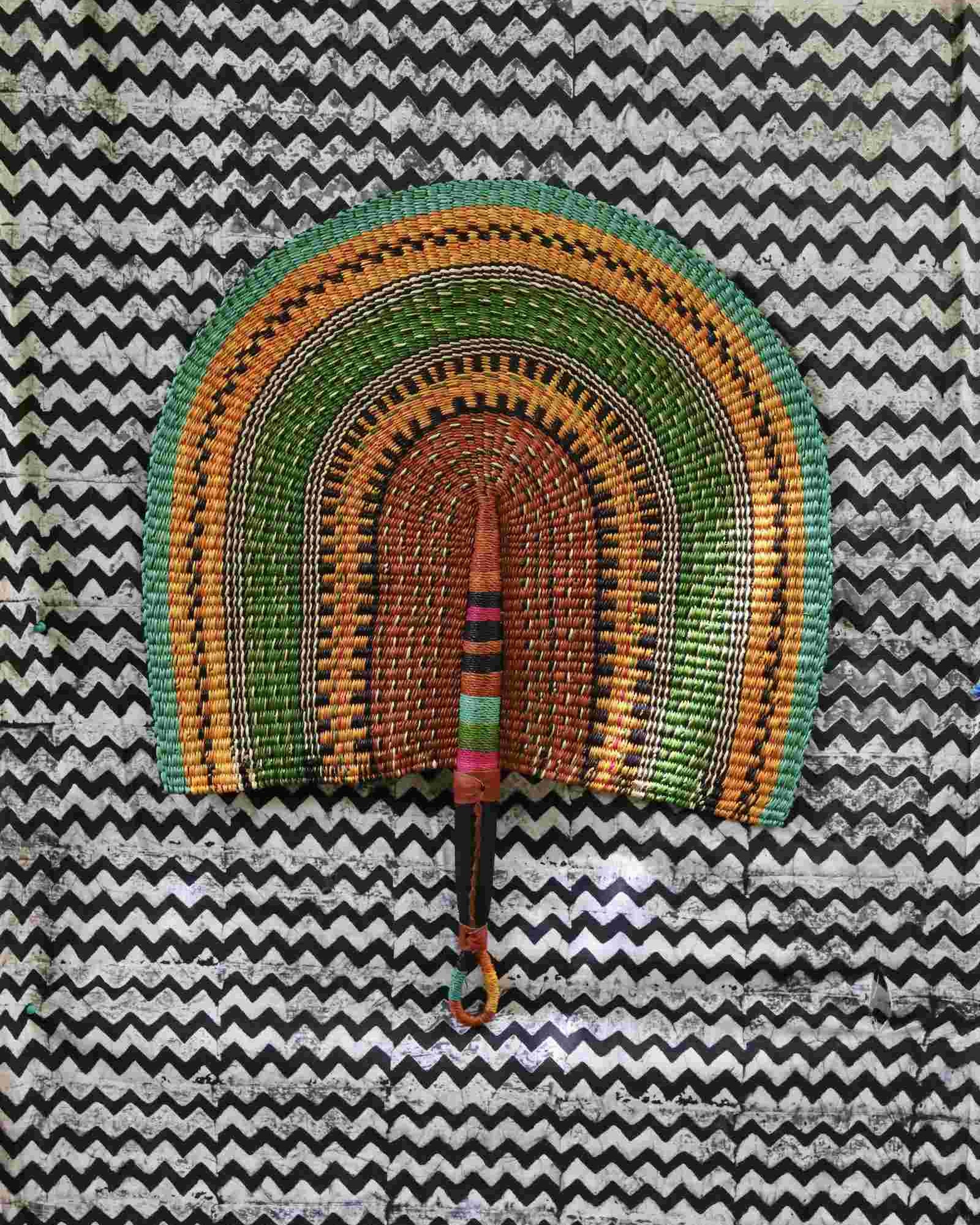 Naba Penga handwoven wall fan with arch bands in green, amber and terracotta, on a black-and-white chevron backdrop.