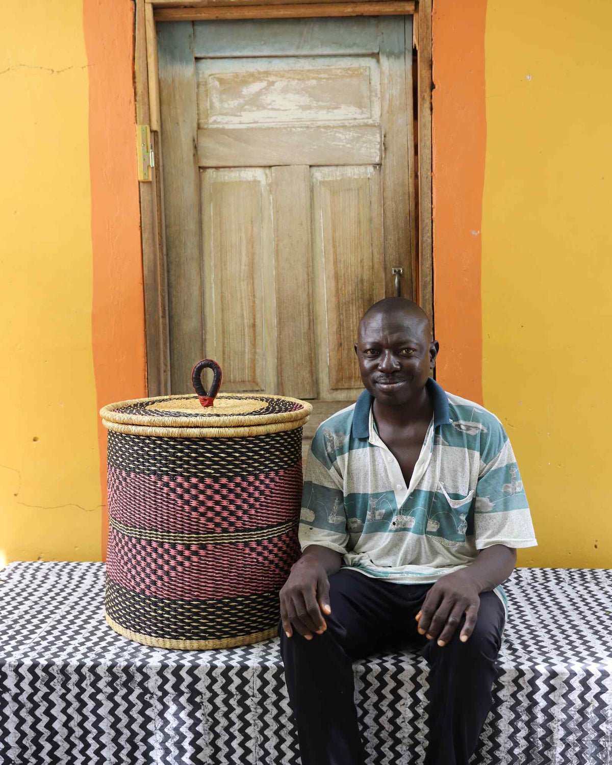 Handwoven Bolga laundry basket in natural with red-pink checkered band, beside a seated man in stripes.