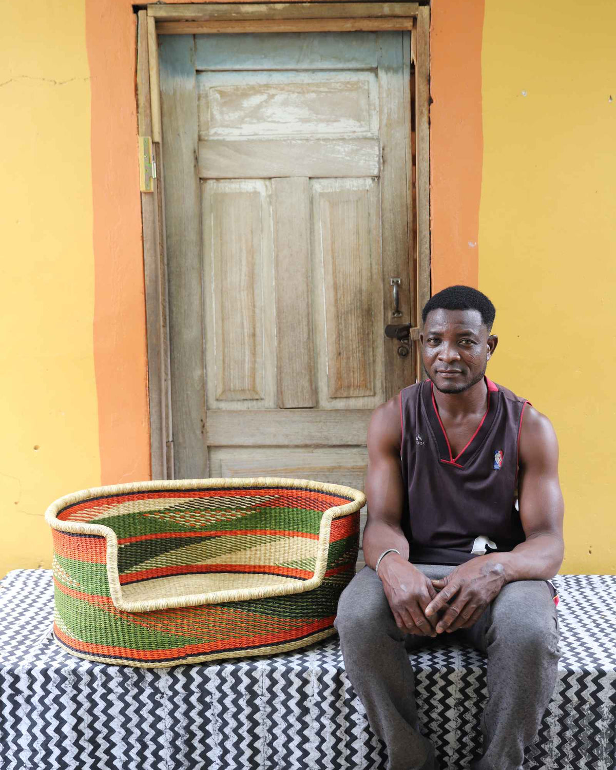 Large handwoven Bolga pet bed in natural straw with green, red and orange bands, beside a seated man.