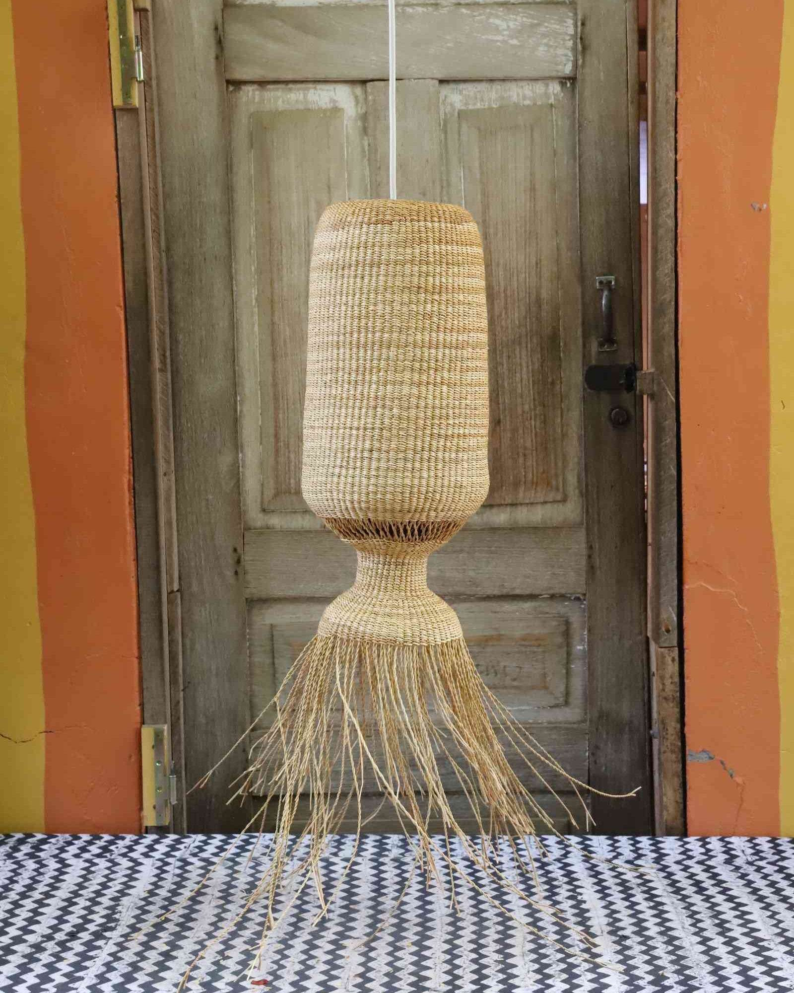 Abyssal pendant lampshade in woven elephant grass with a fringed base, suspended before a weathered wooden door.