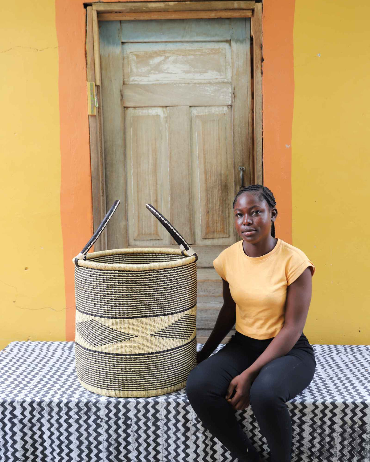 Large handwoven Bolga laundry basket in natural with dark diamond motif, beside a seated woman in yellow.