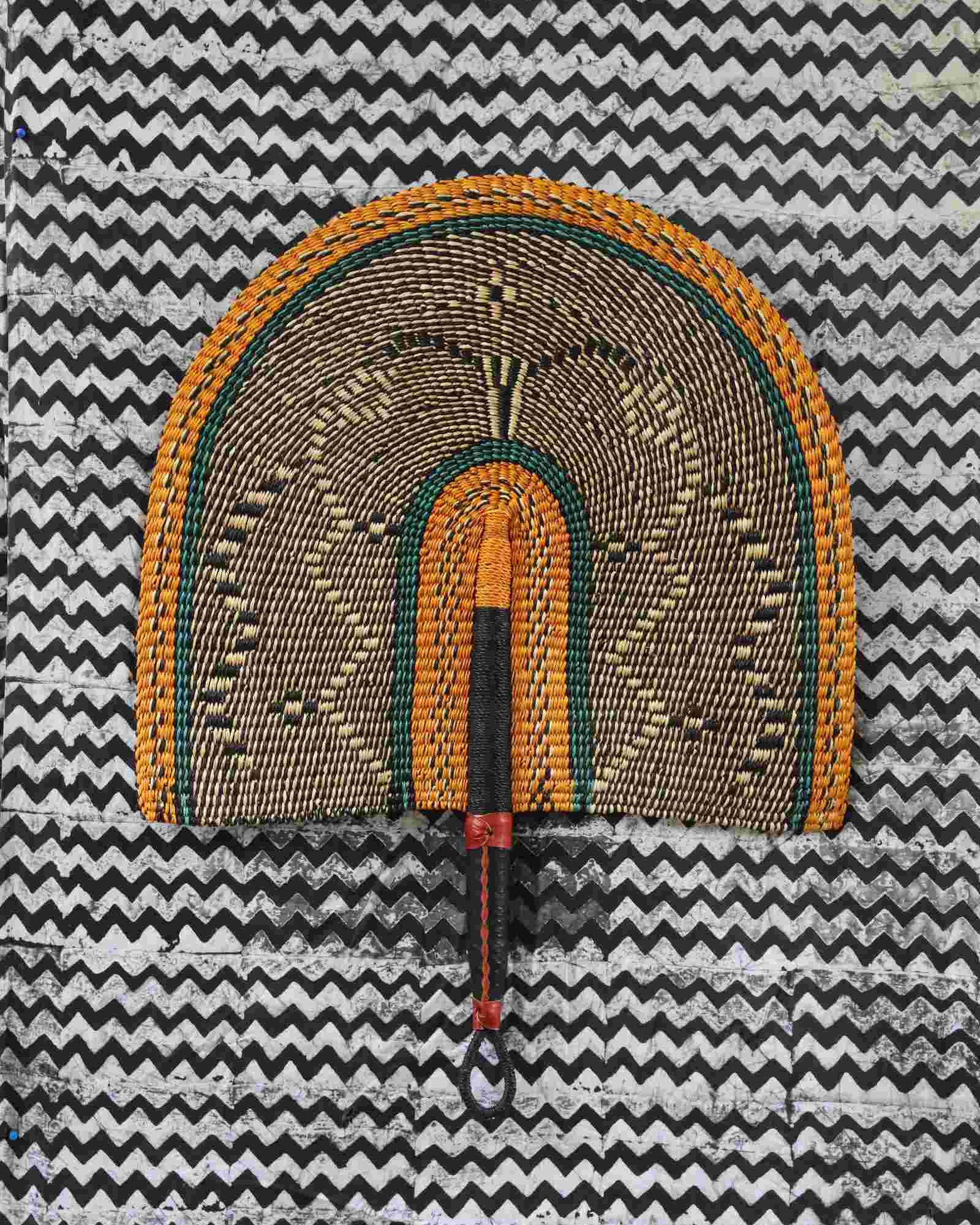 Naba Penga wall fan in natural straw with a mustard border and green trim, laid on a chevron cloth.