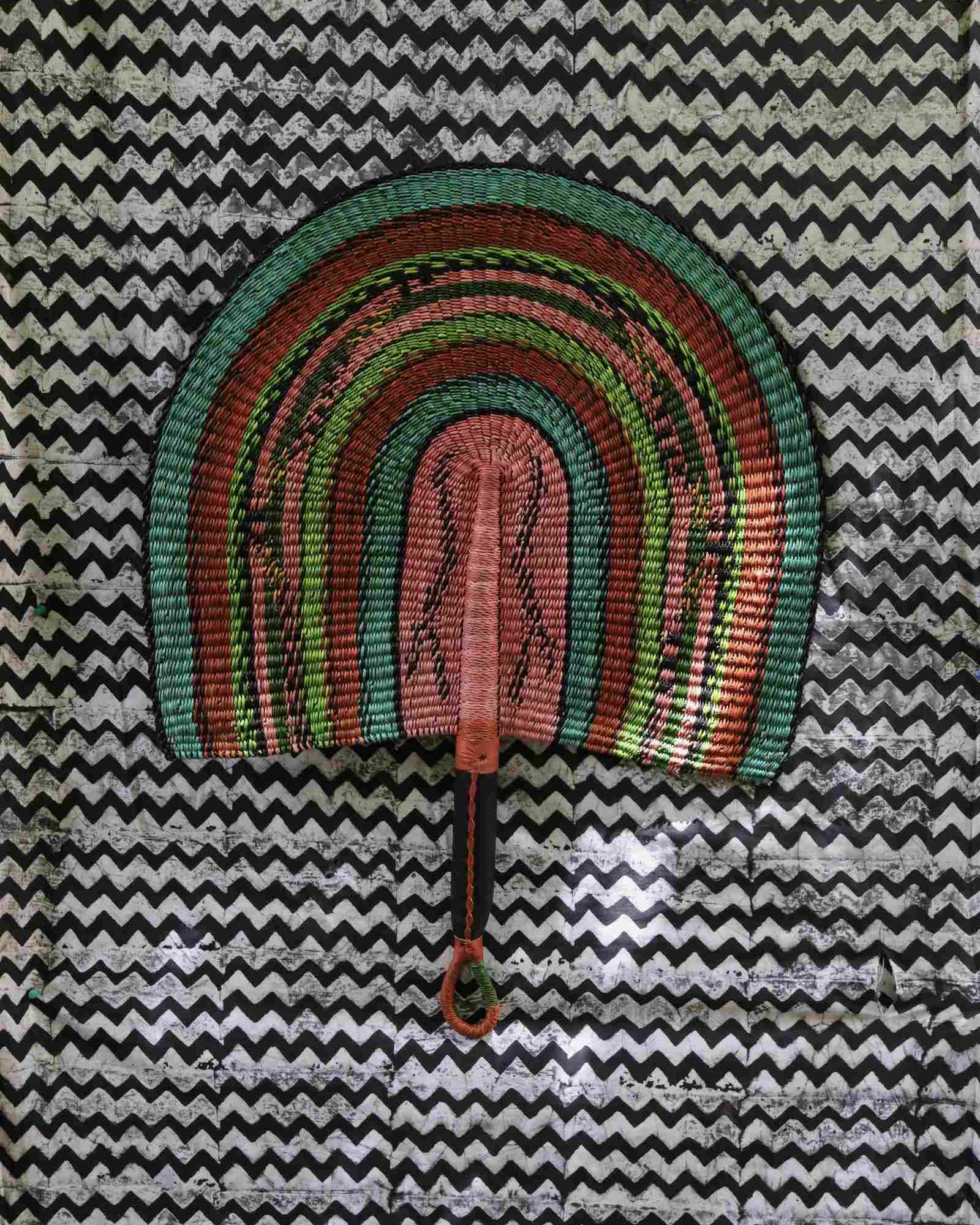 Naba Penga handwoven wall fan in teal, terracotta and dusty pink banded stripes, on a chevron-print backdrop.