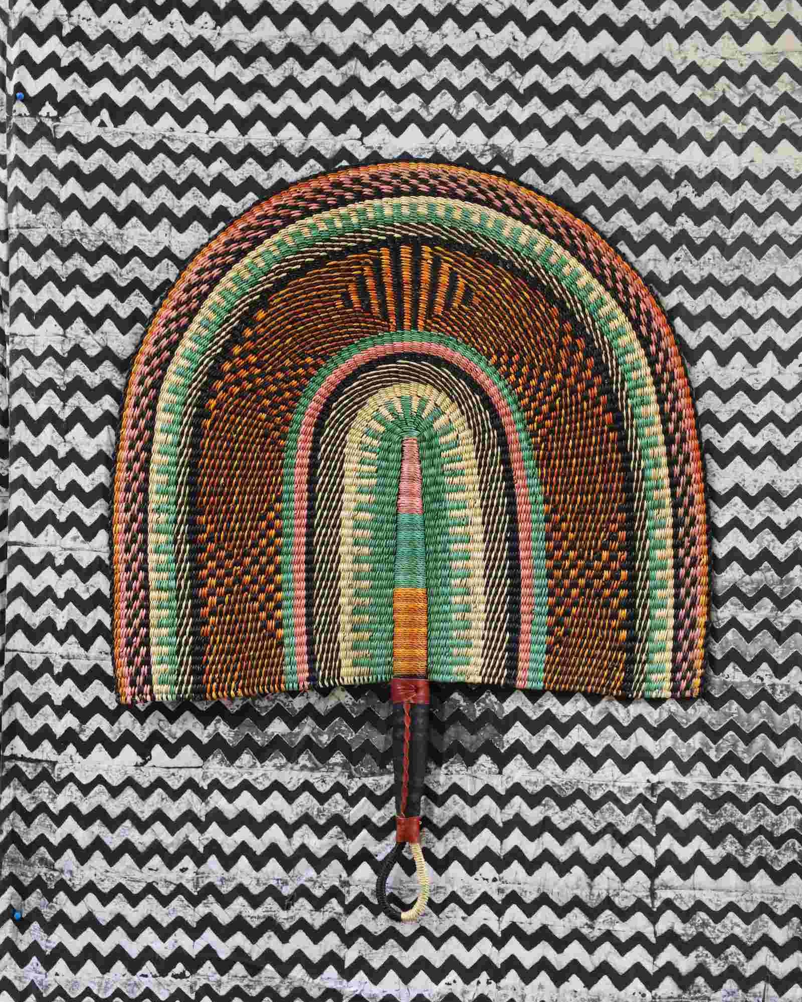 Naba Penga wall fan in natural straw with sage green and rust banded arches, laid on a chevron cloth.