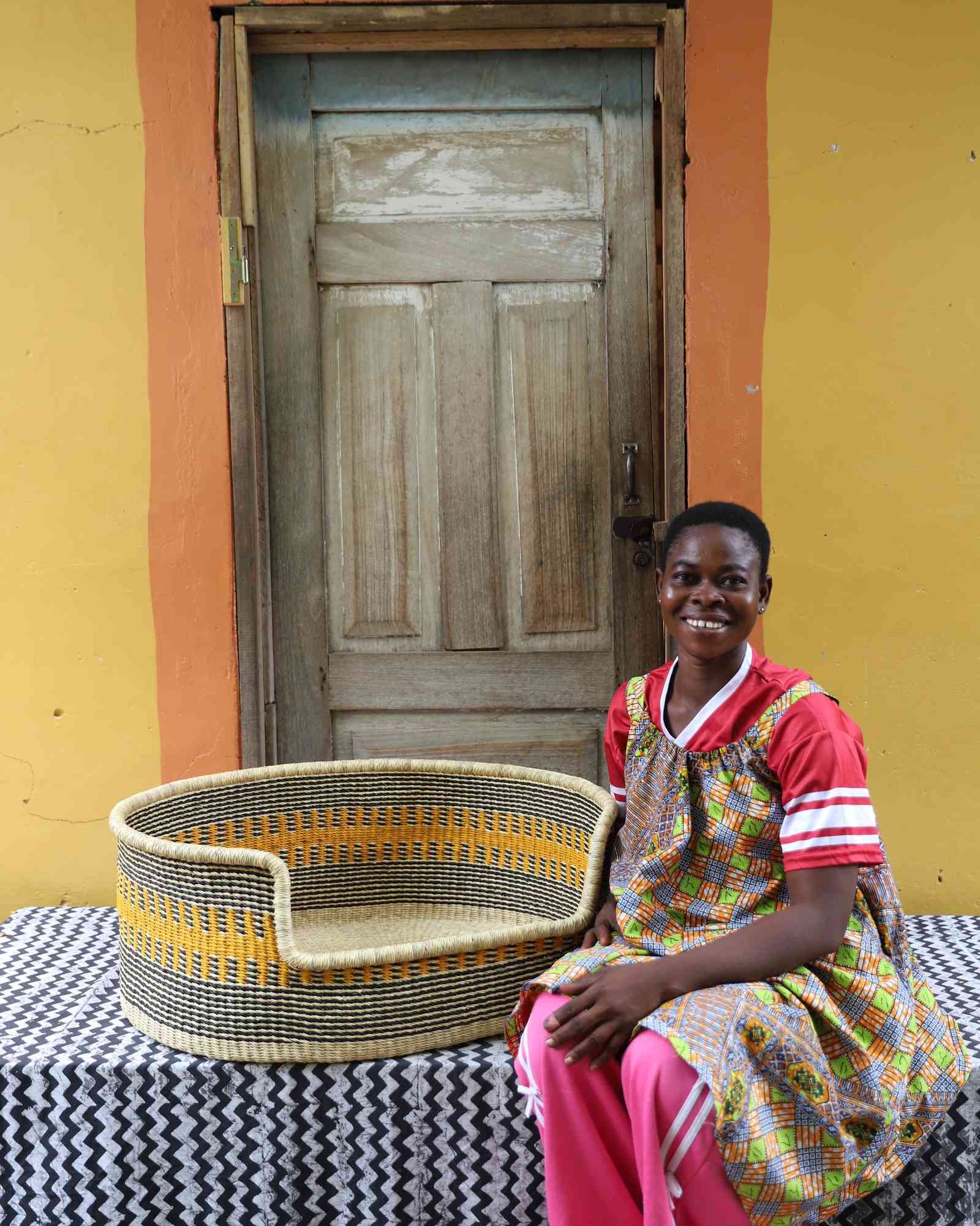 Large handwoven Bolga pet bed in natural straw with orange and yellow bands, beside a seated woman in red.