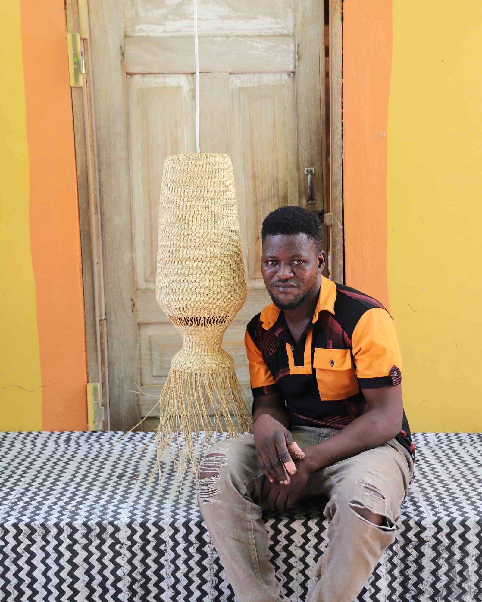 Man seated on a woven stool beneath the Abyssal lampshade in a weathered wooden doorway.