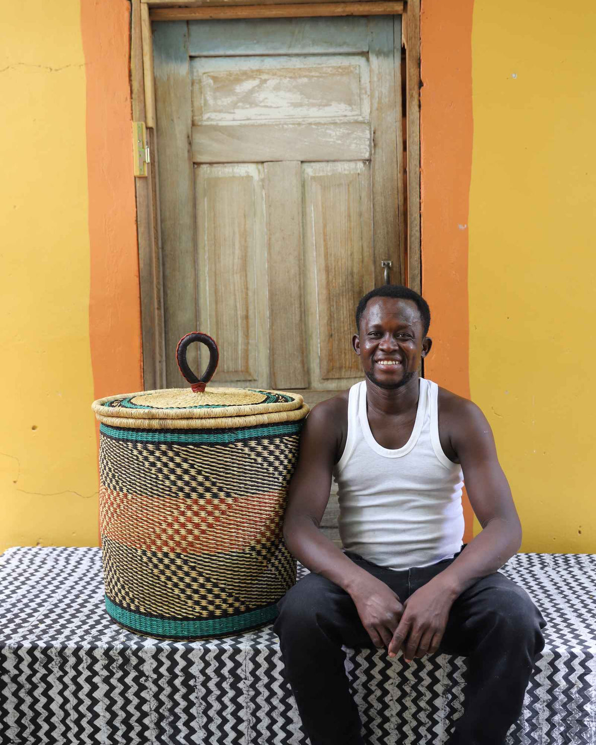 Handwoven Bolga laundry basket in natural with orange and dark bands, beside a seated man in a white vest.