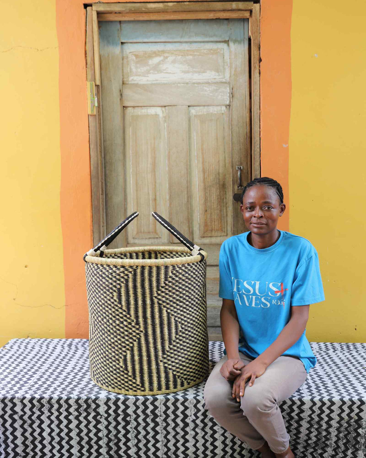 Large handwoven Bolga laundry basket in natural with fine black diamond weave, beside a seated woman in blue.