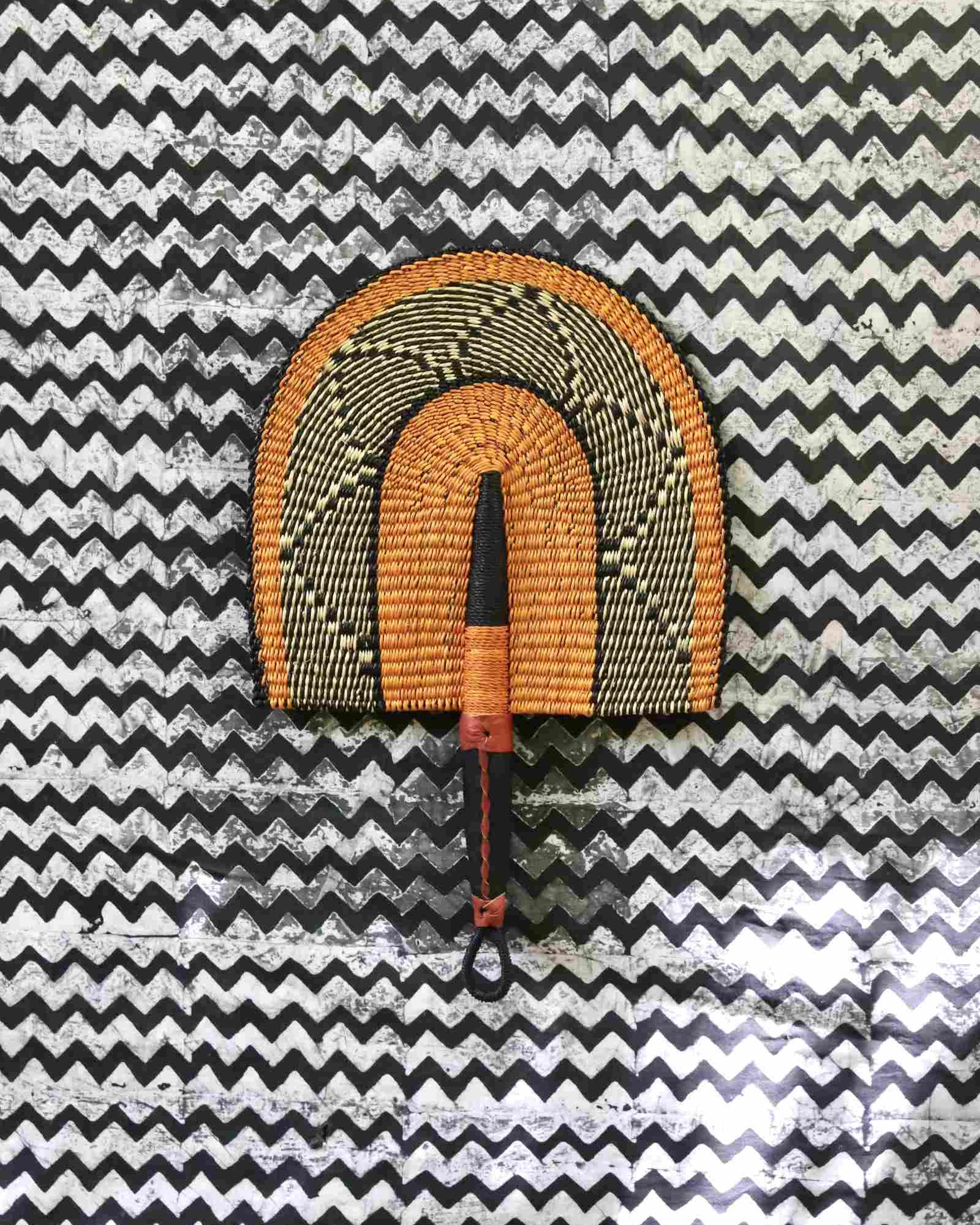 Handwoven Bolga Fan in natural straw with black geometric outer arch, on a black-and-white chevron fabric backdrop.