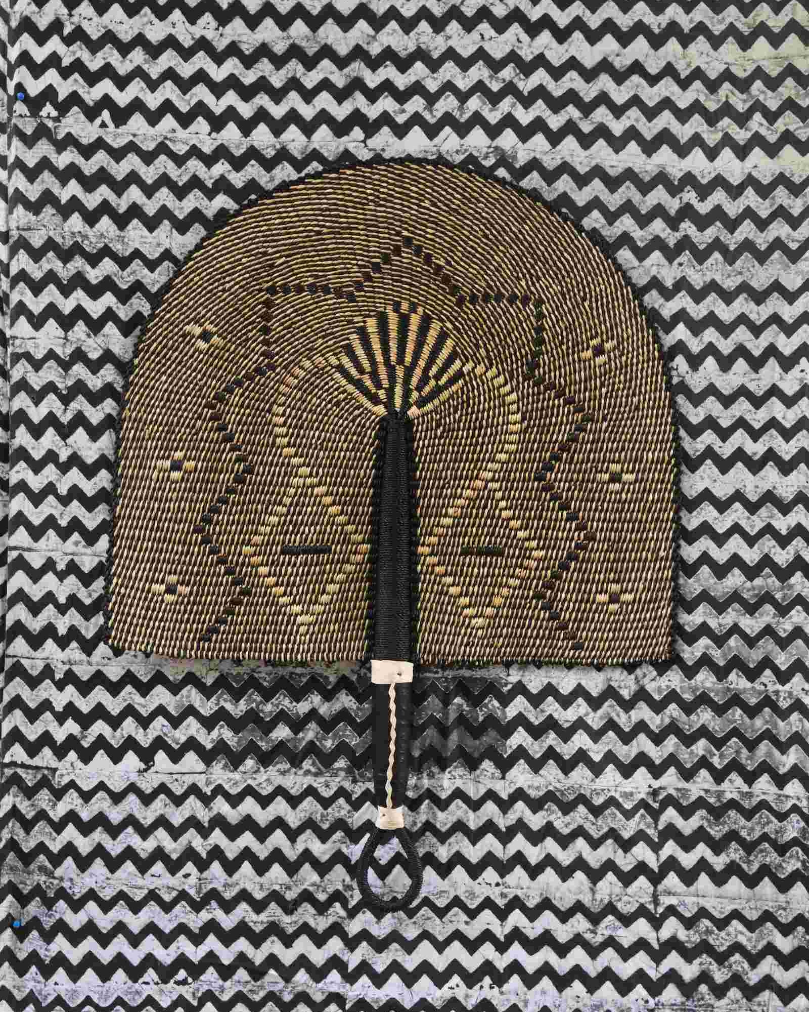 Naba Penga wall fan in dark brown straw with a tan star-shaped geometric weave, laid on a chevron cloth.