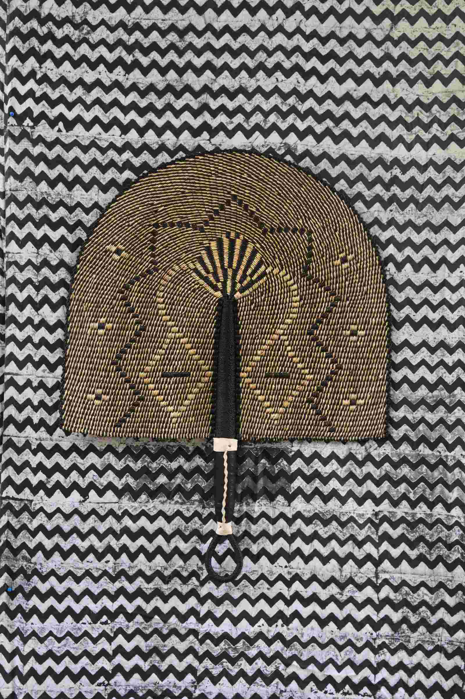 Naba Penga wall fan in dark brown straw with a tan star-shaped geometric weave, laid on a chevron cloth.
