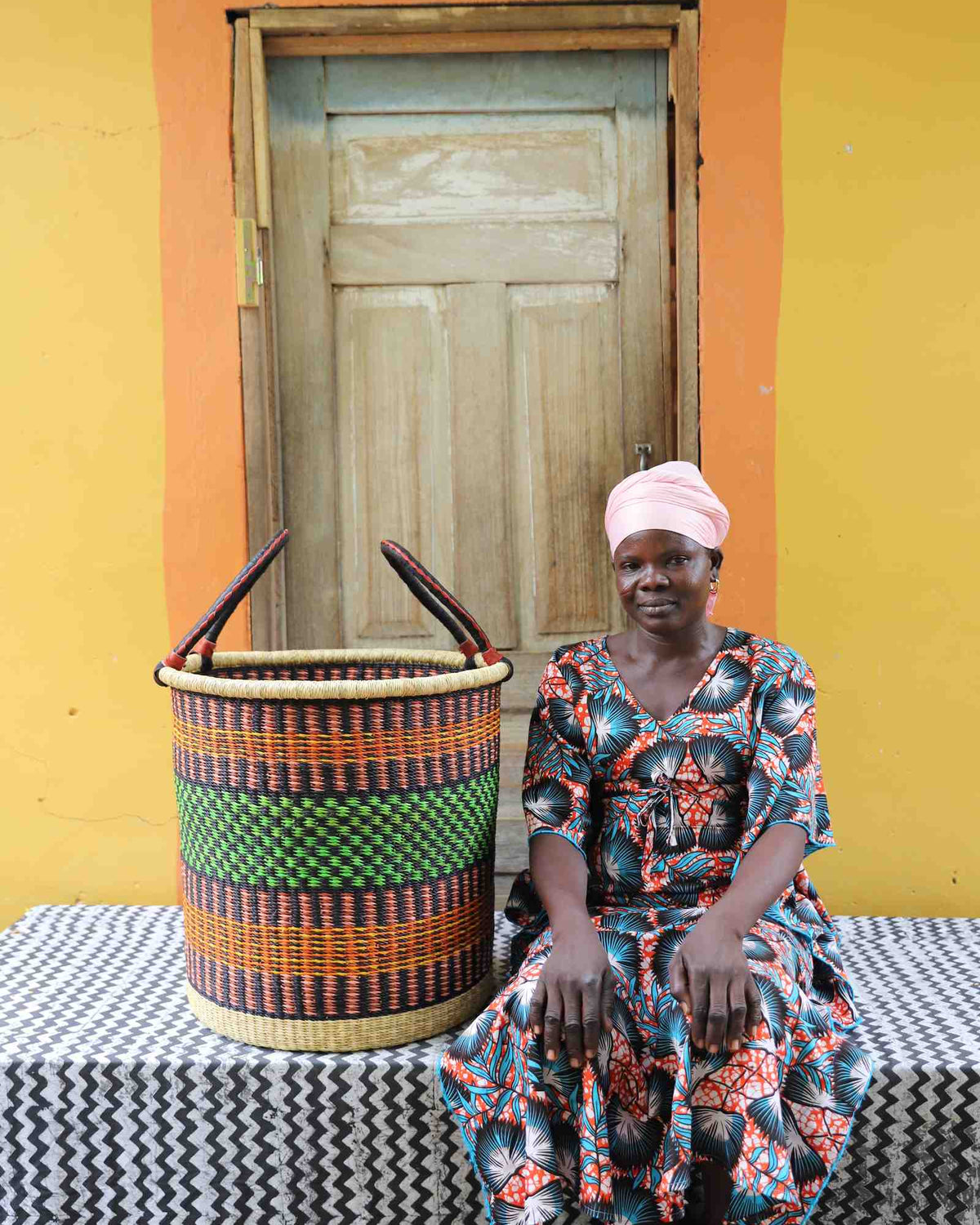 Large handwoven Bolga laundry basket in dark straw with green and orange bands, beside a seated woman in floral.