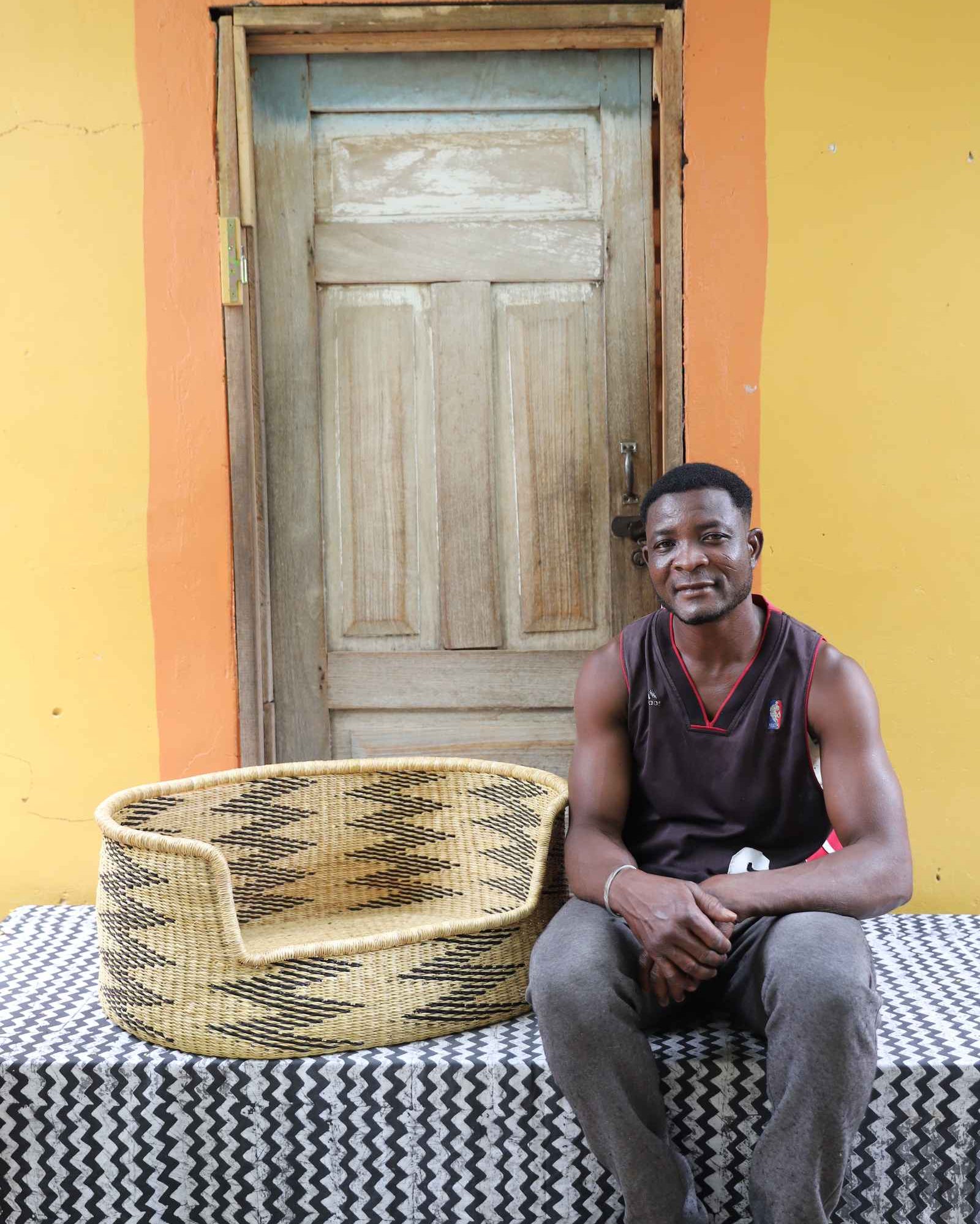 Large handwoven Bolga pet bed in natural straw with black arrows, beside a seated man in a dark vest.