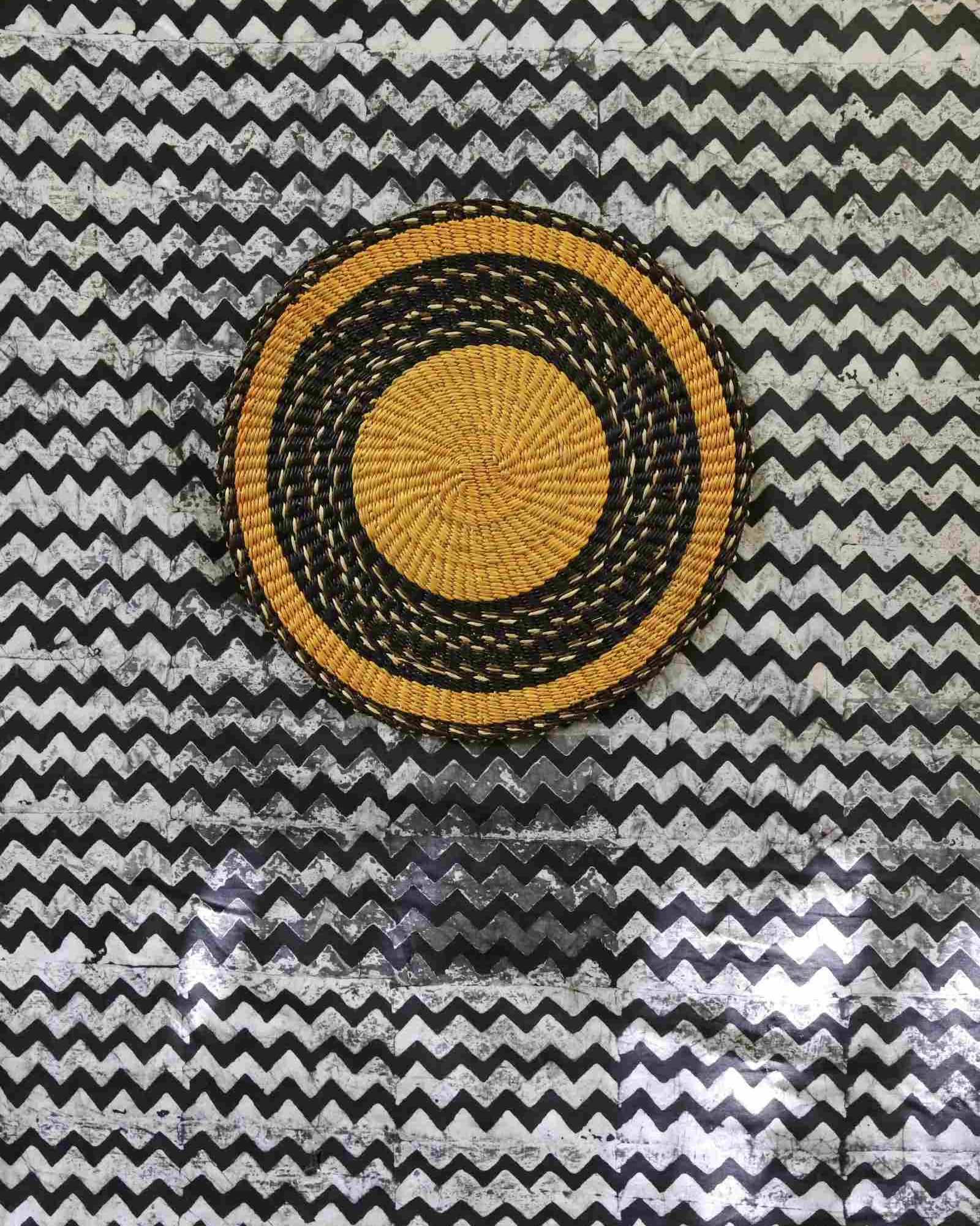 Handwoven Bolga placemat in natural straw with bold black border ring, on a black-and-white chevron fabric backdrop.