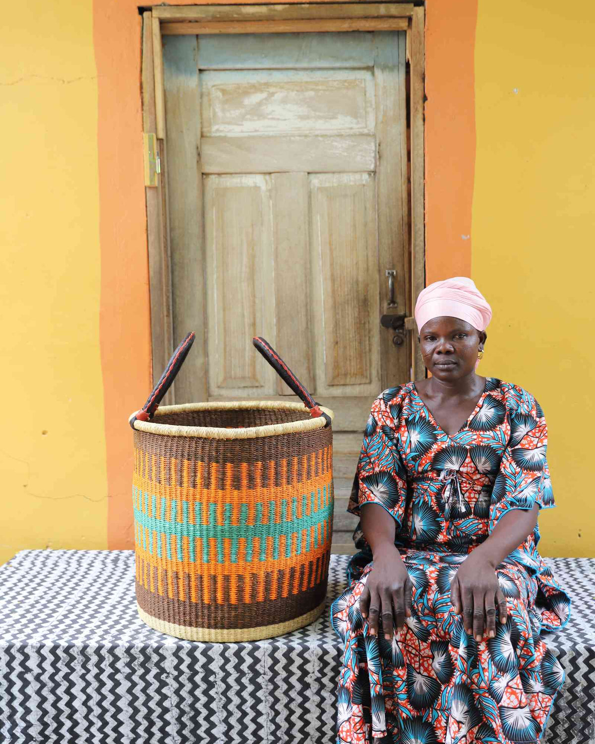 Large handwoven Bolga laundry basket in brown and orange bands, beside a seated woman in a printed dress.