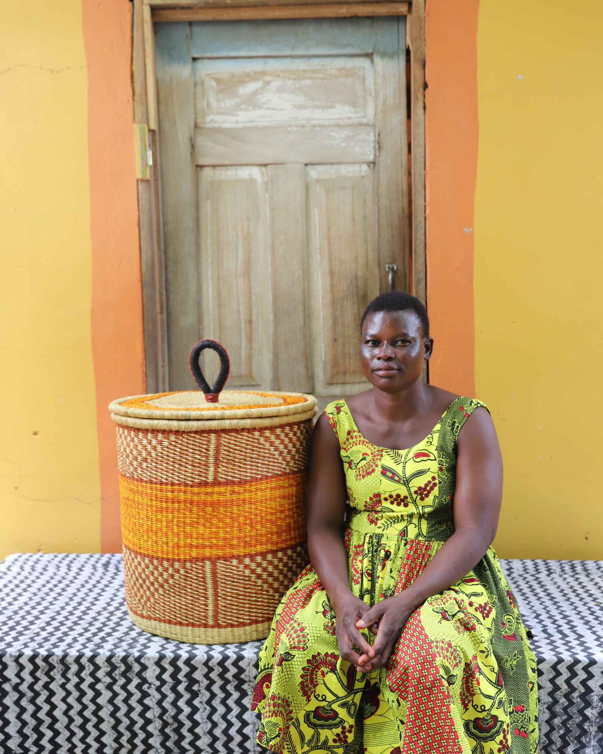 Handwoven Bolga laundry basket with orange central band, beside a seated woman in green and yellow.