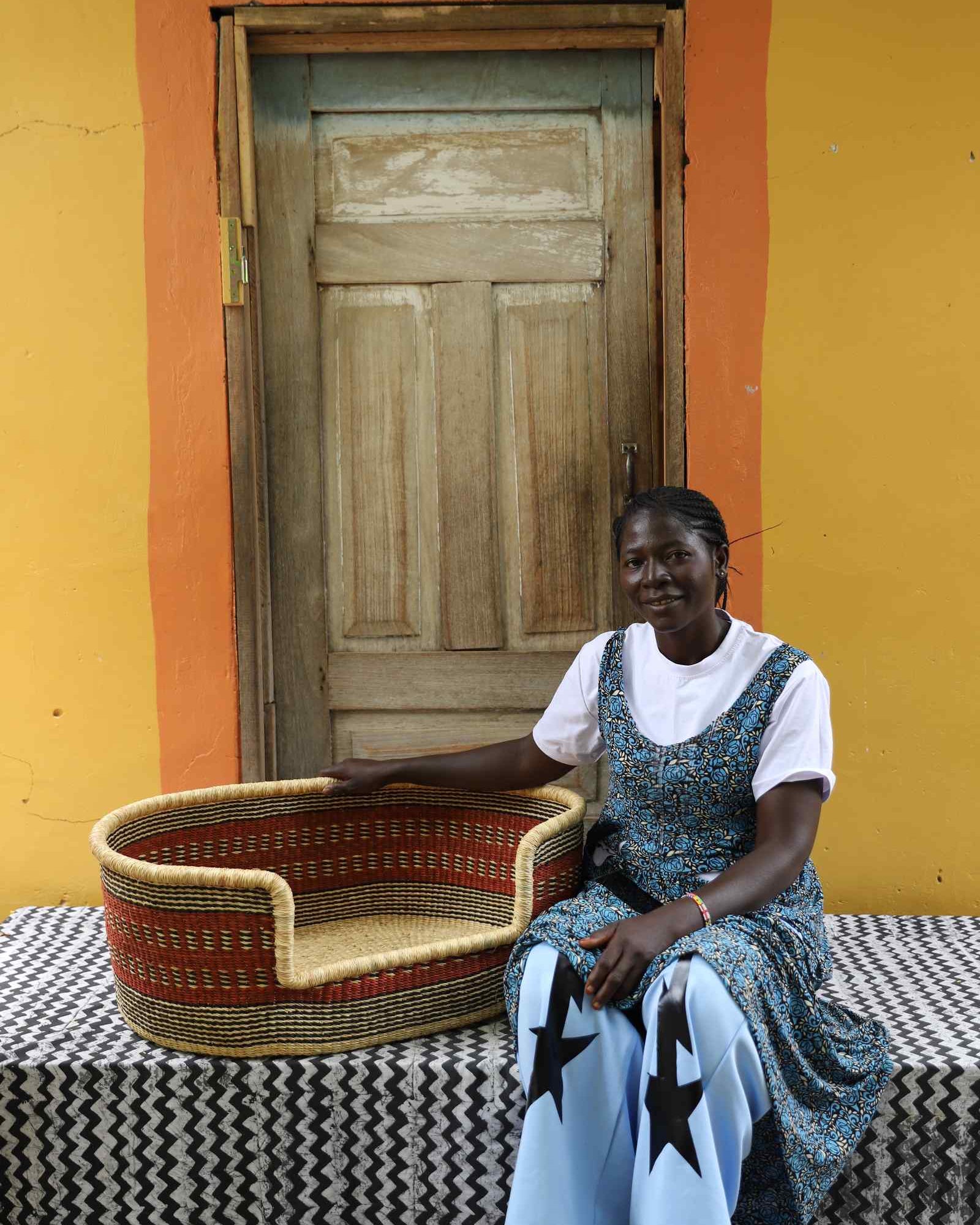 Medium handwoven Bolga pet bed in natural straw with red bands, beside a seated woman in a printed dress.