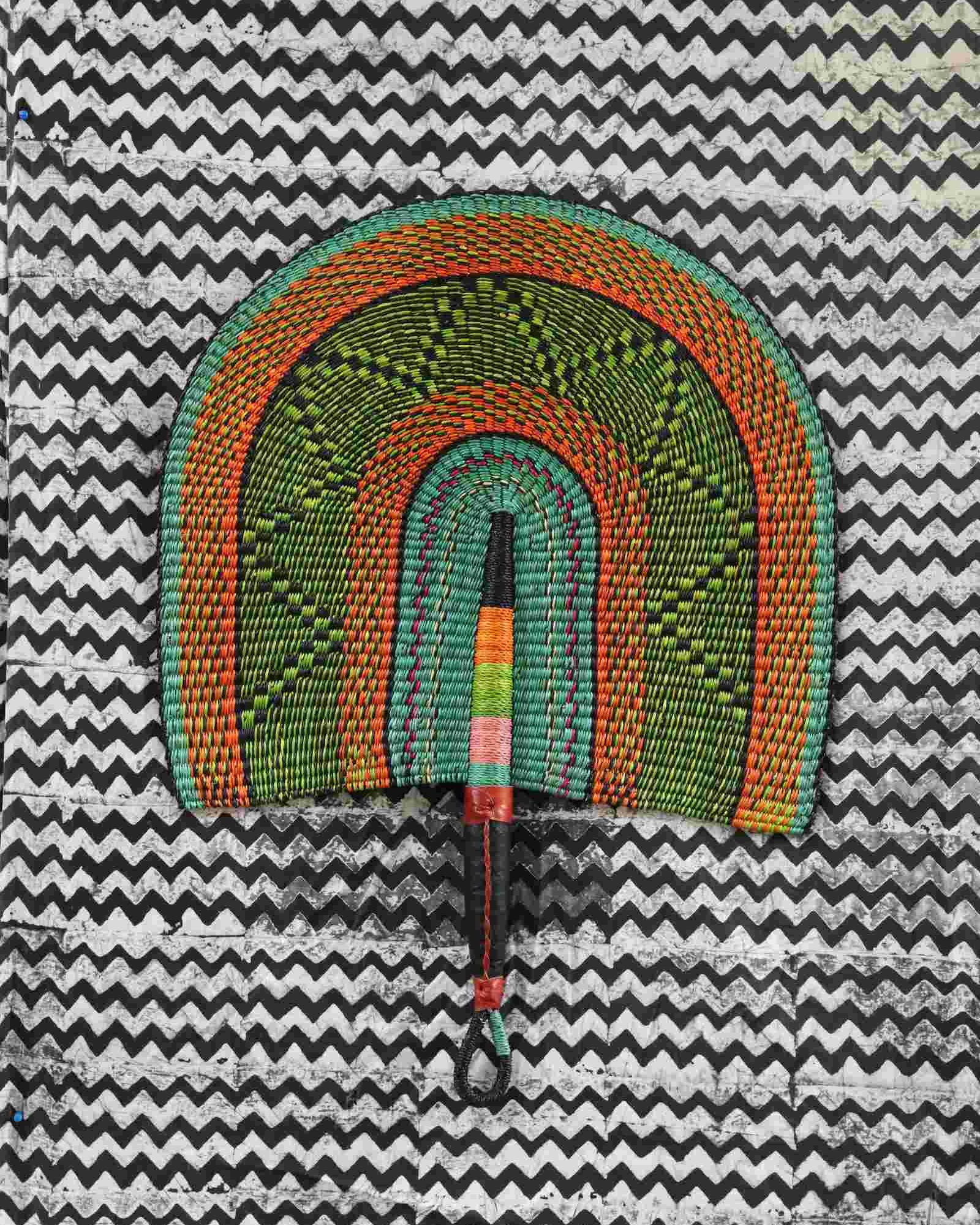 Naba Penga wall fan in green woven straw with an orange border and diamond pattern, laid on a chevron cloth.