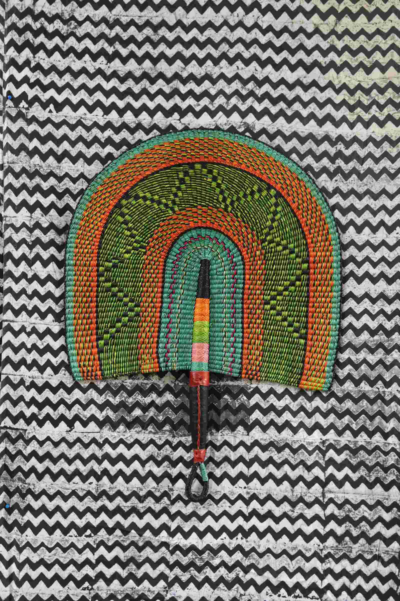 Naba Penga wall fan in green woven straw with an orange border and diamond pattern, laid on a chevron cloth.