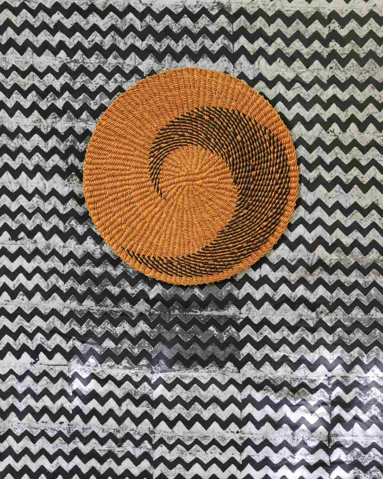 Handwoven Bolga placemat in natural straw with black spiral pattern, on a black-and-white chevron fabric backdrop.