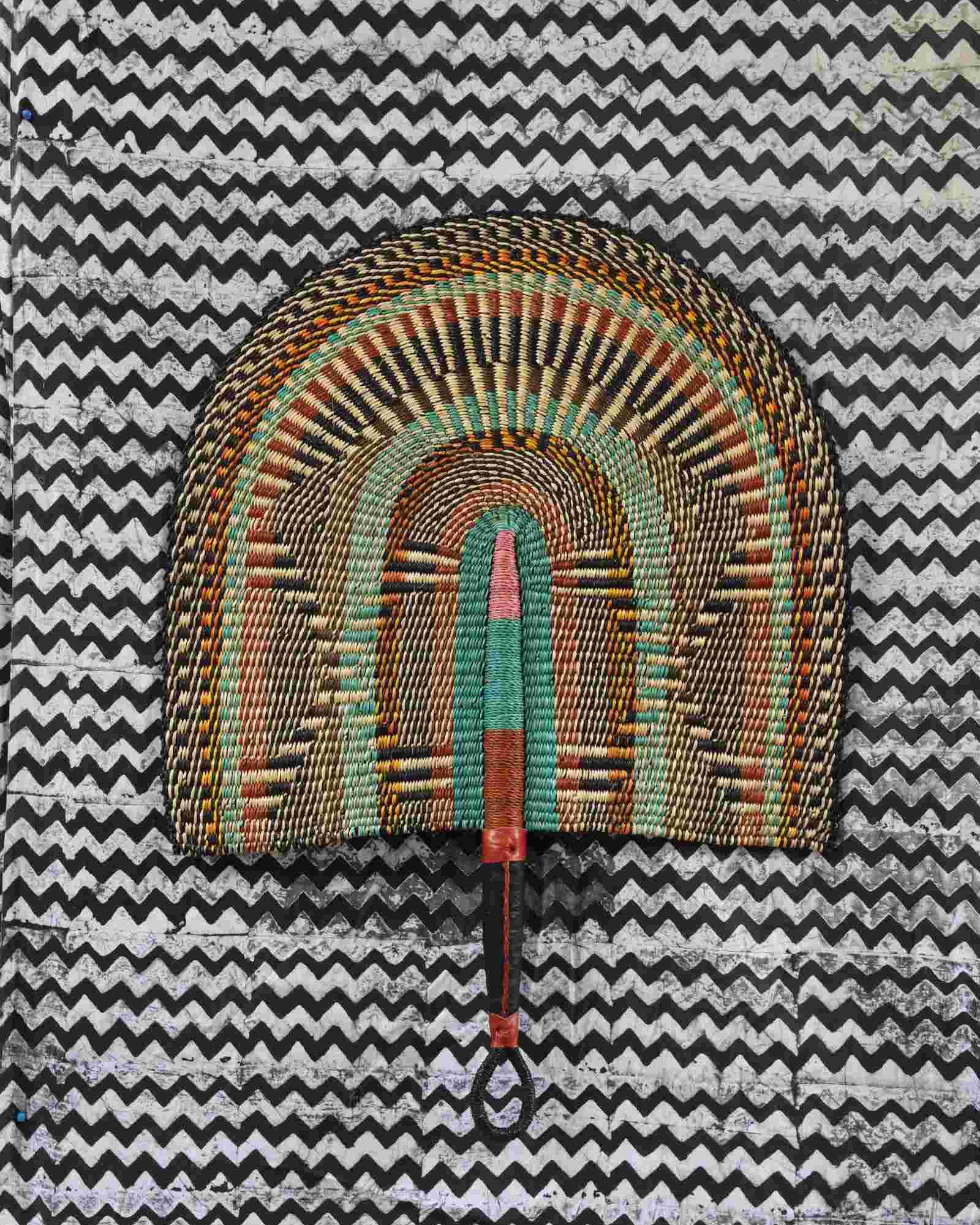 Naba Penga wall fan in natural straw with rust, green and teal concentric arches, laid on a chevron cloth.