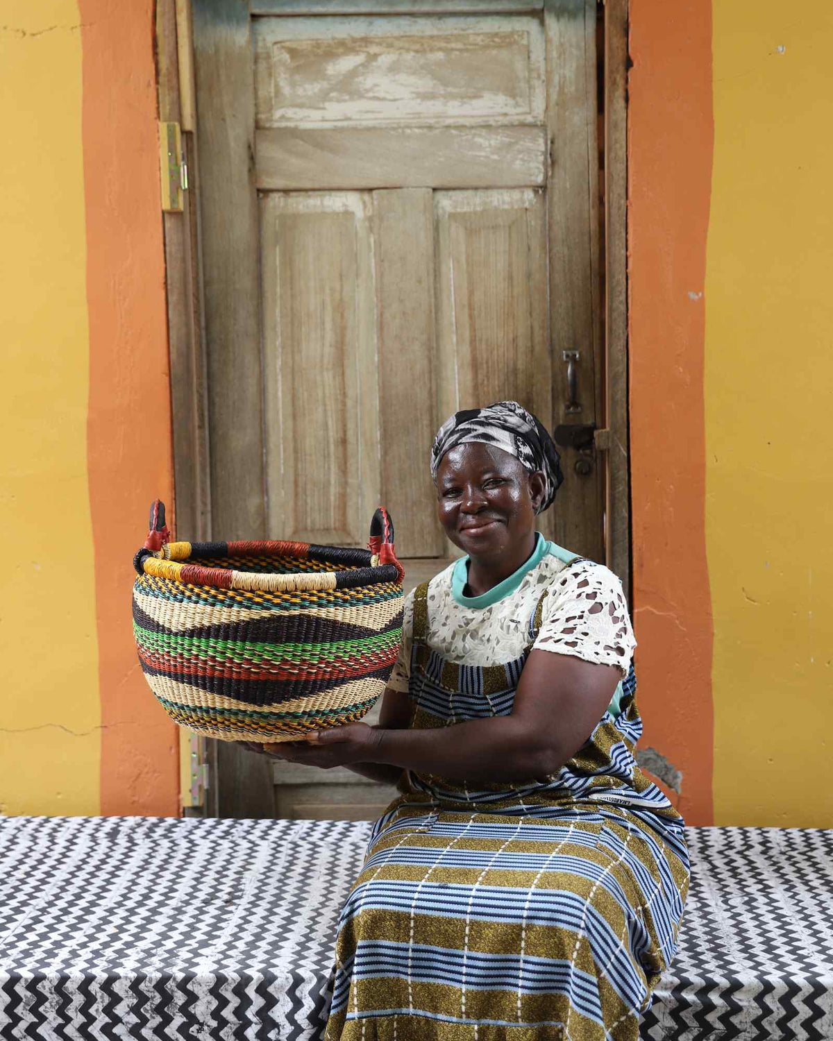 Small handwoven Bolga Gbenka storage basket in multicolour bands, styled beside a seated woman in olive.