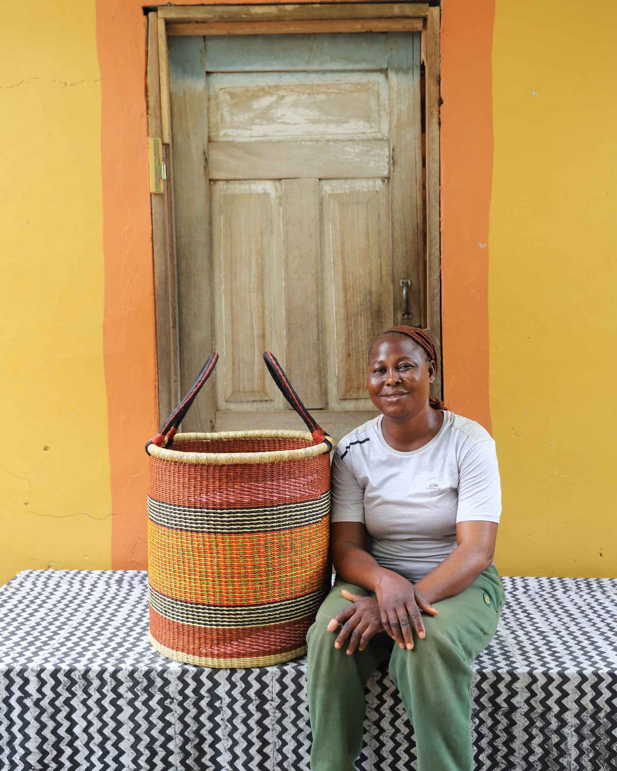 Medium handwoven Bolga laundry basket in orange, yellow and red bands, beside a seated woman in olive.